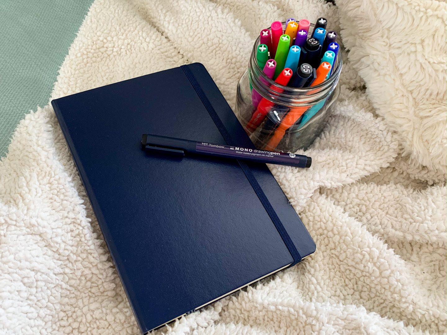 a flatlay that consists of a white fuzzy blanket, with a navy blue bullet journal laid on top, along with one pen laid on top of the journal, and a jar full of colourful pens sitting beside it - one of my purhcases in my low buy year