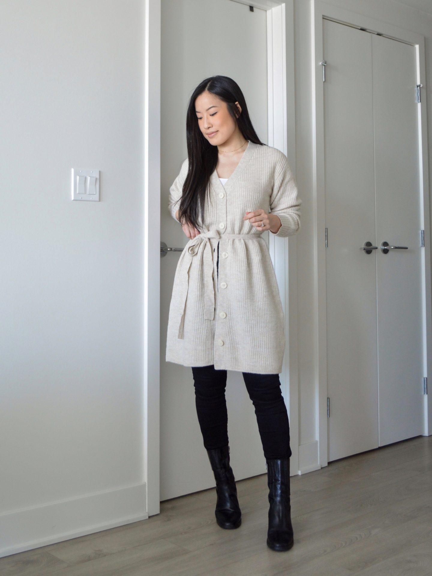 Sharon of Her Simple Sole is wearing the Frank and Oak The Recycled Wool Cardigan Dress buttoned halfway down with a white tank top, black skinny jeans and black ankle boots.