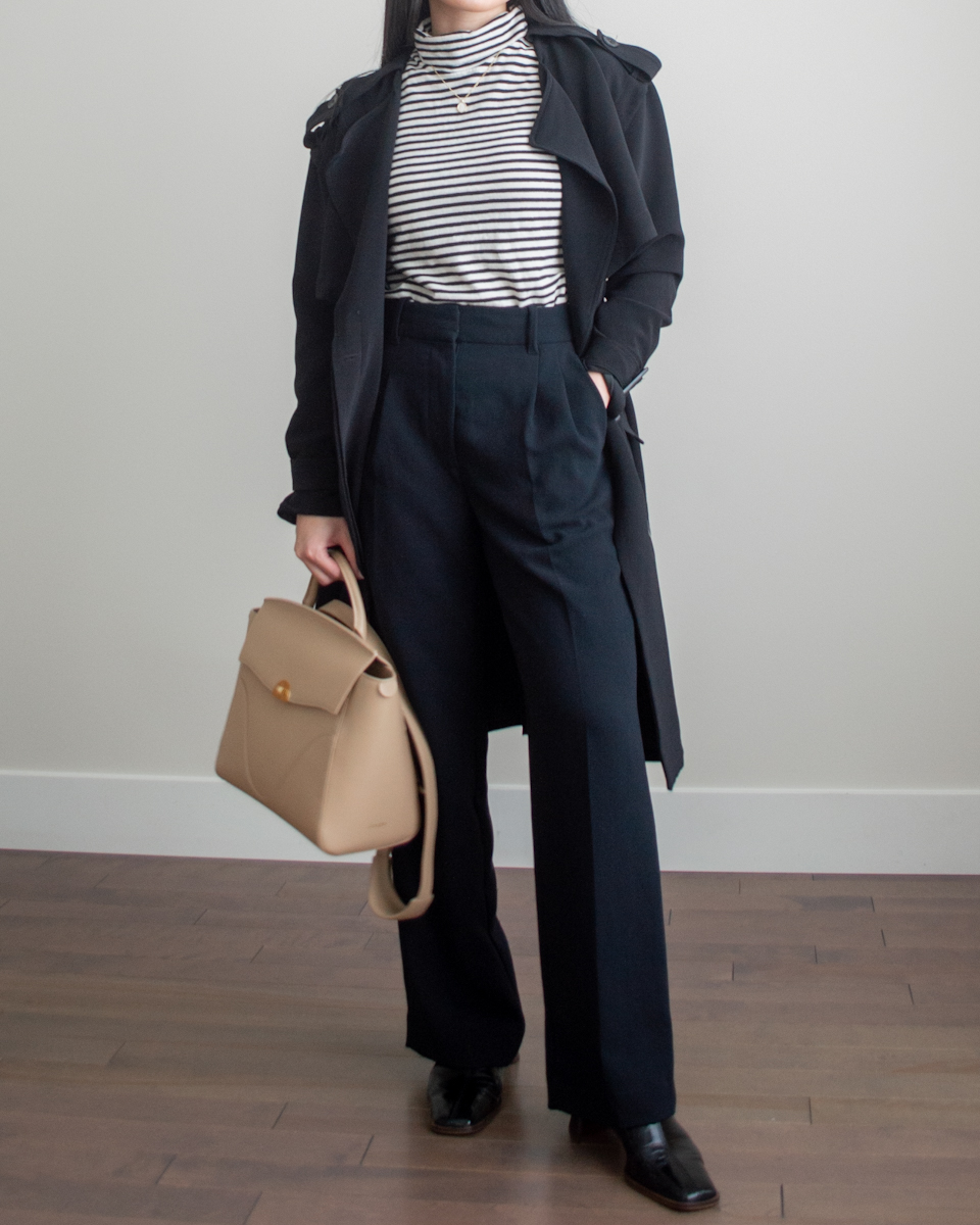 Her Simple Sole outfit photo featuring a black trench coat, stripe turtleneck top, black wide leg trousers, black heeled mules, and the Oleada Wavia Bag in Champagne