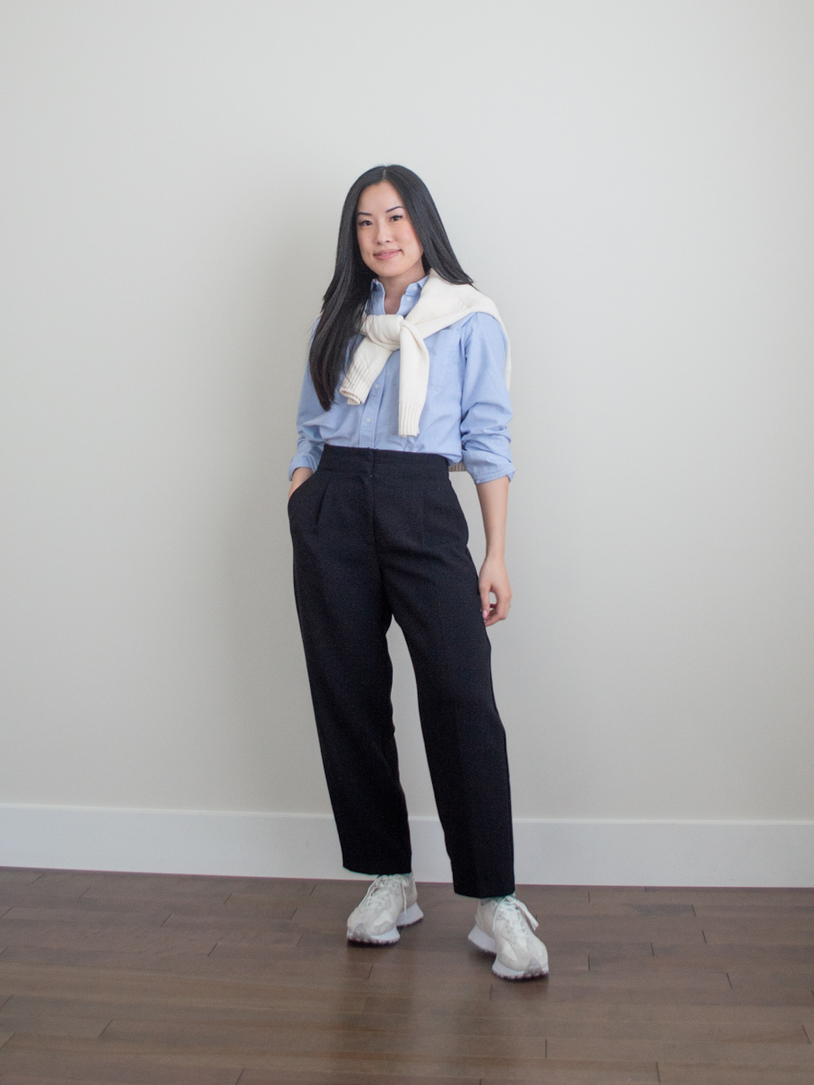 Her Simple Sole outfit photo featuring blue relaxed oxford shirt, cream cotton sweater draped over shoulders, black straight leg trousers, New Balance sneakers