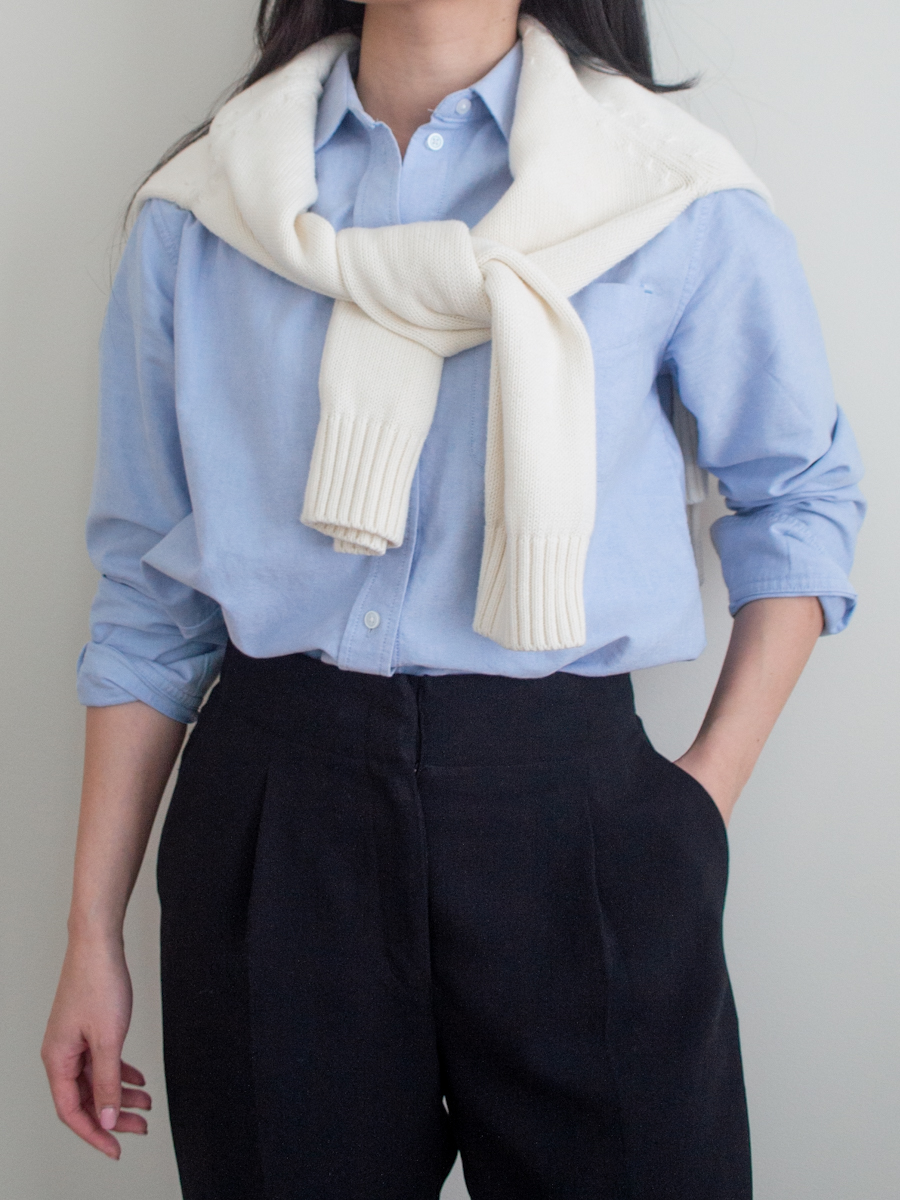 close up outfit photo of a blue relaxed oxford shirt styled with a cream sweater over the shoulders and black trousers, intentional gifts under $100