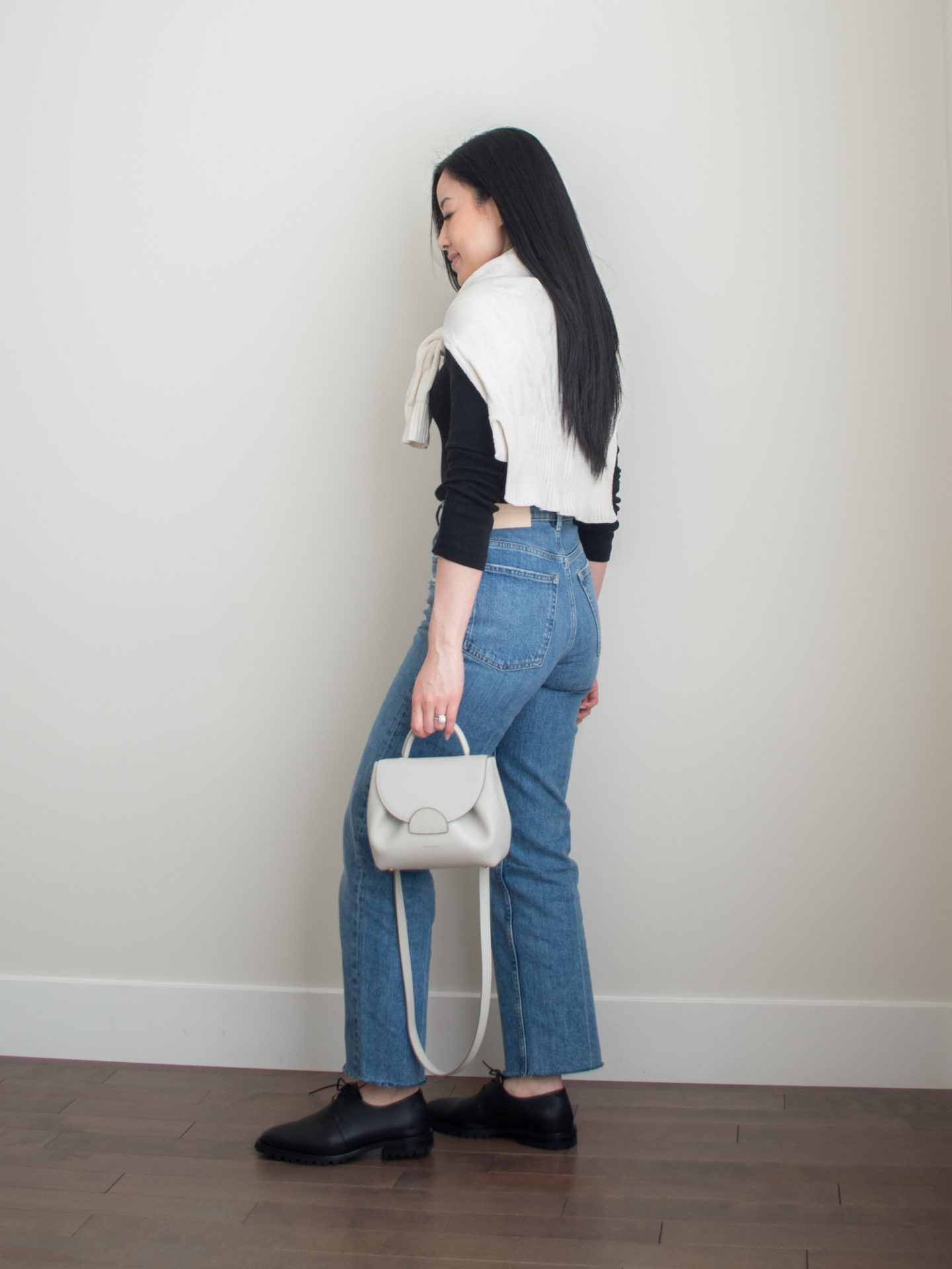 Smart casual outfit idea: basic black fitted long sleeve top, cream sweater over the shoulders, blue straight leg jeans, black oxford shoes, cream crossbody bag