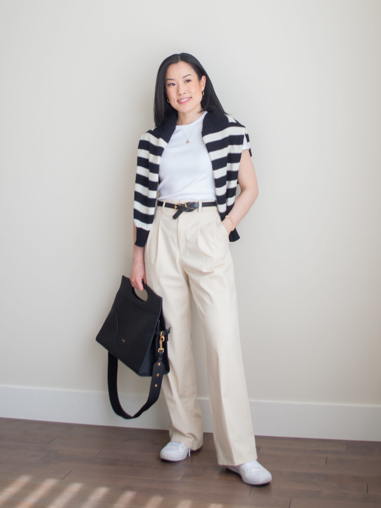 A casual chic outfit worn by Sharon of Her Simple Sole, featuring a black and white stripe sweater over the shoulders, a white ribbed crop t-shirt, cream wide leg pants, monochrome white Converse Chuck Taylor sneakers, gold dainty jewelry, and the Oleada Captain Briefcase in Caviar | casual chic style | minimalist outfit | spring outfit idea