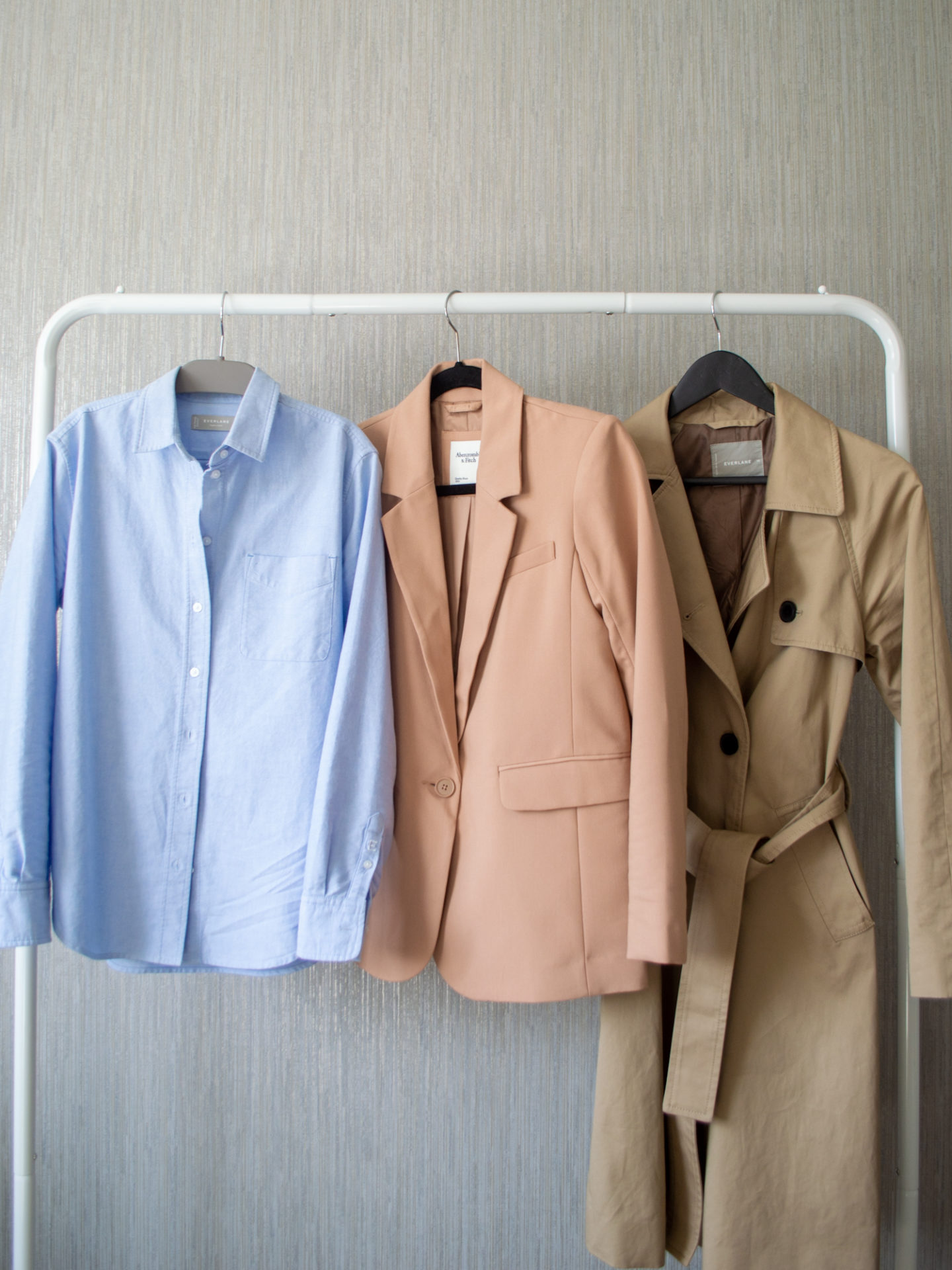 Intentional Life and Style Content Creator Sharon of Her Simple Sole showcases her spring smart casual capsule wardrobe picks for layering pieces on a hanging rack, including a classic brown trench coat, a warm brown classic blazer, and a blue relaxed oxford button up shirt