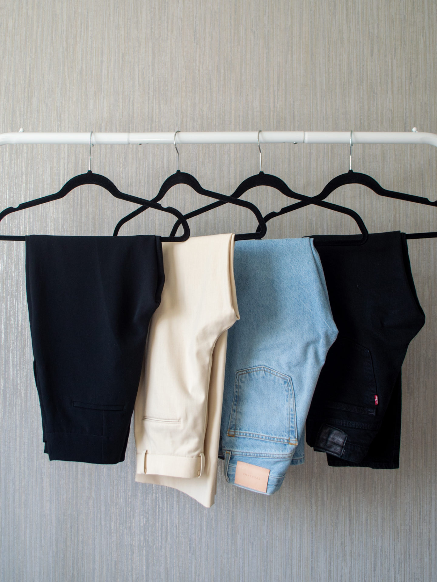 Intentional Life and Style Content Creator Sharon of Her Simple Sole showcases her spring smart casual capsule wardrobe picks for bottoms on a hanging rack, including black straight leg jeans, light wash blue straight fit jeans, cream wide leg pants, and black straight leg pants