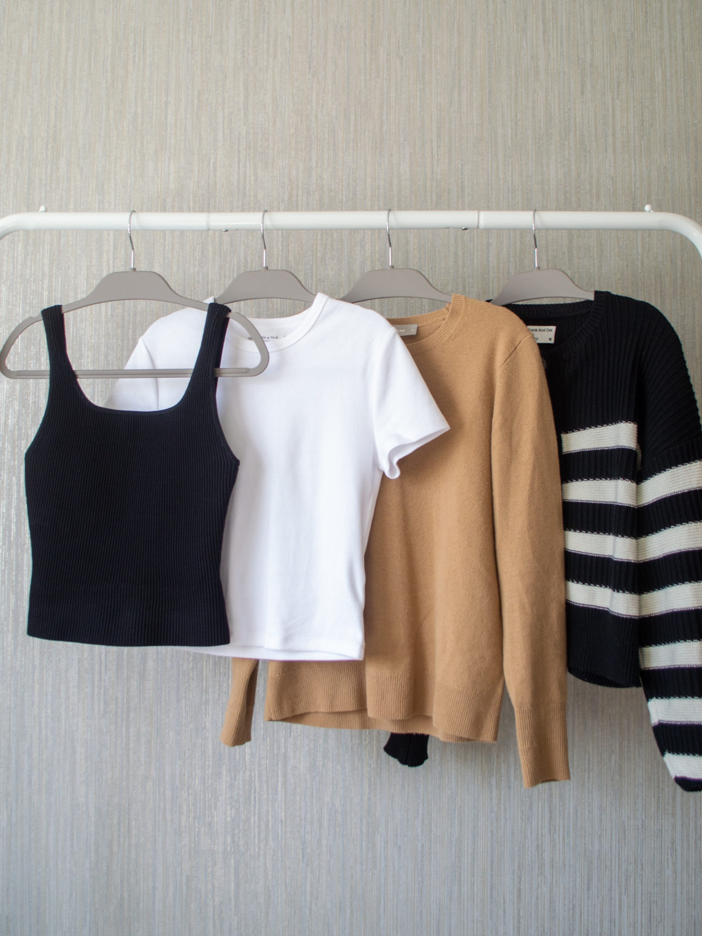 Intentional Life and Style Content Creator Sharon of Her Simple Sole showcases her spring smart casual capsule wardrobe picks for tops on a hanging rack, including a black and white stripe cardigan, a camel cashmere crewneck sweater, a white ribbed baby t-shirt, and a black ribbed square neck tank top