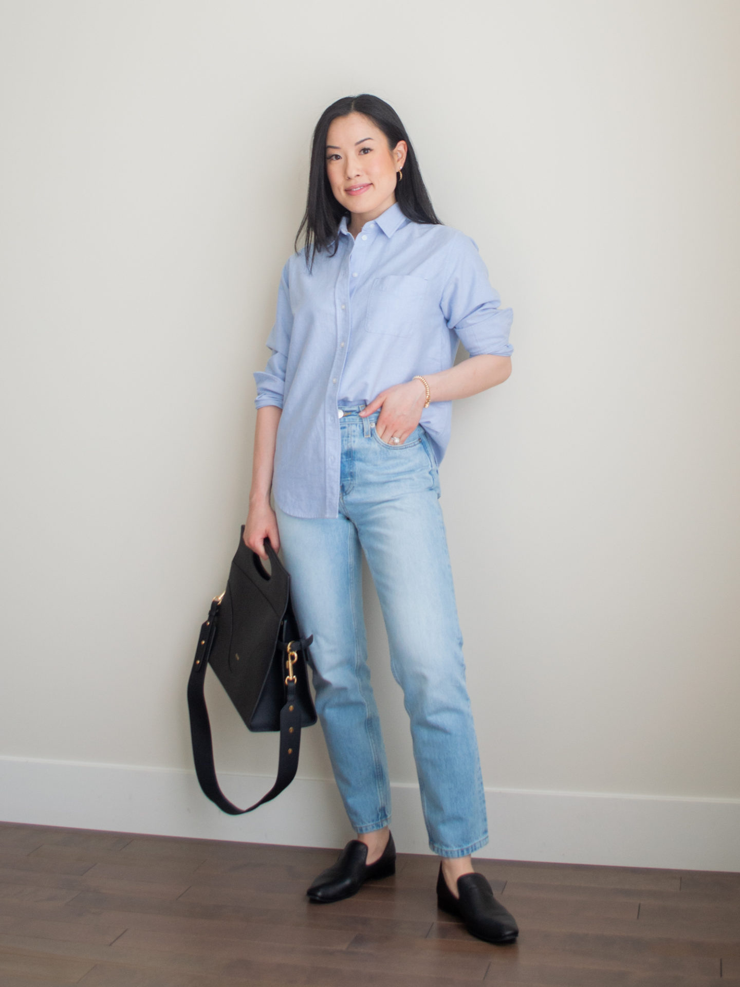 A casual chic outfit worn by Sharon of Her Simple Sole featuring a blue relaxed oxford shirt, blue straight leg jeans, classic black loafers, gold dainty jewelry, and the Oleada Captain Briefcase bag in Caviar | blue monochrome outfit | minimalist outfit | spring outfit idea