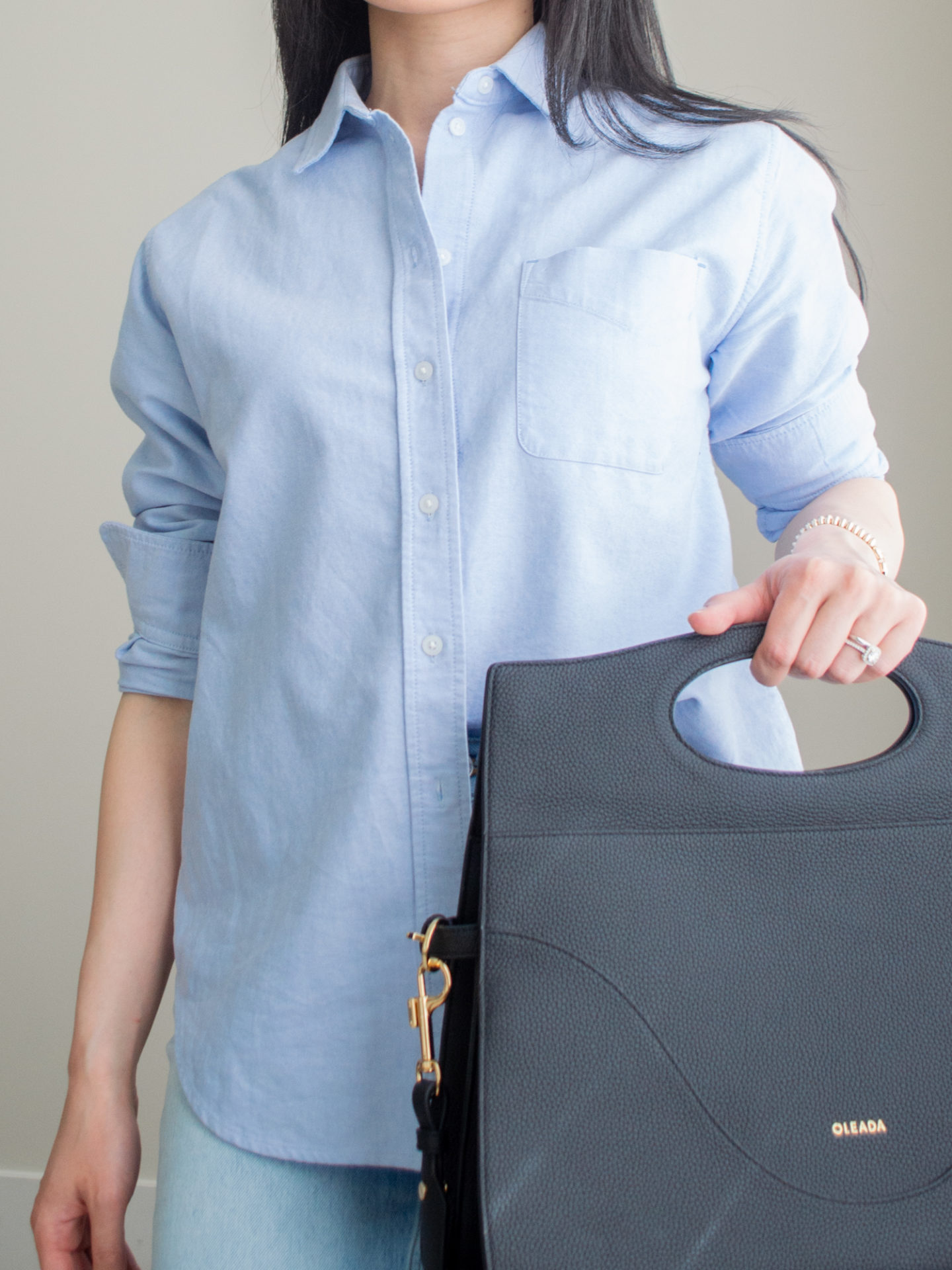 Close up shot of a casual chic outfit worn by Sharon of Her Simple Sole featuring a blue relaxed oxford shirt, blue straight leg jeans, gold dainty jewelry, and the Oleada Captain Briefcase bag in Caviar | blue monochrome outfit | minimalist outfit | spring outfit idea