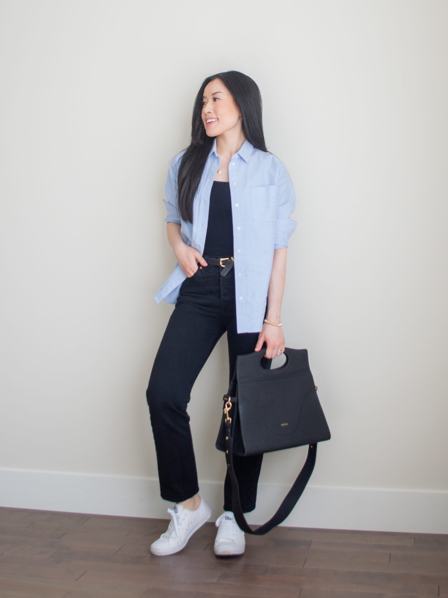 A casual chic outfit worn by Sharon of Her Simple Sole featuring a blue relaxed oxford shirt, black straight leg jeans, monochrome white Converse Chuck Taylor sneakers, a ribbed square neck tank top, gold dainty jewelry, and the Oleada Captain Briefcase bag in Caviar | casual outfit idea | minimalist outfit | spring outfit idea