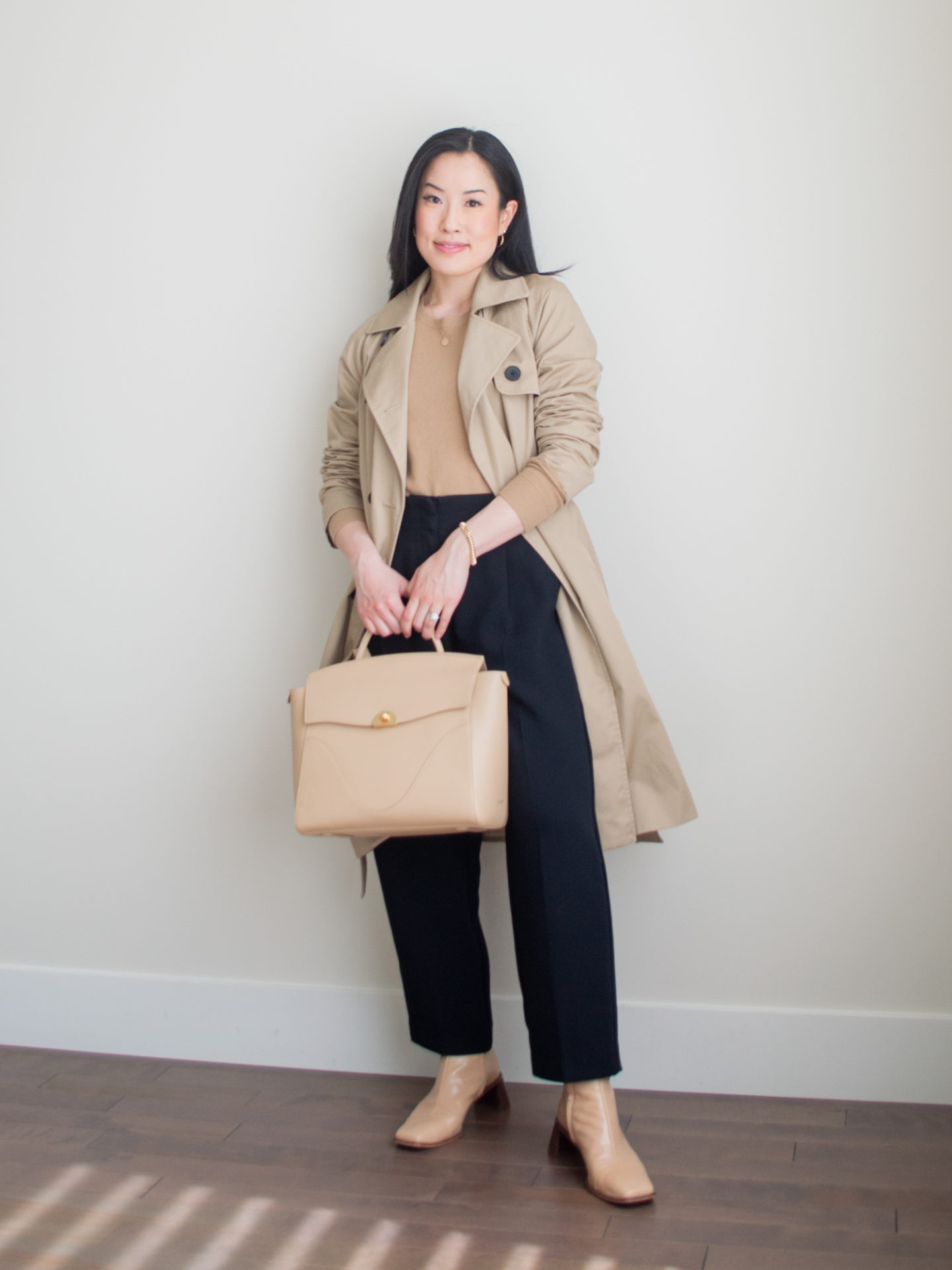 A casual chic outfit worn by Sharon of Her Simple Sole, featuring a camel cashmere crewneck sweater, black straight leg pants, a classic brown trench coat, nude ankle boots, gold dainty jewelry, and the Oleada Wavia bag in Champagne | classic spring outfit | workwear outfits