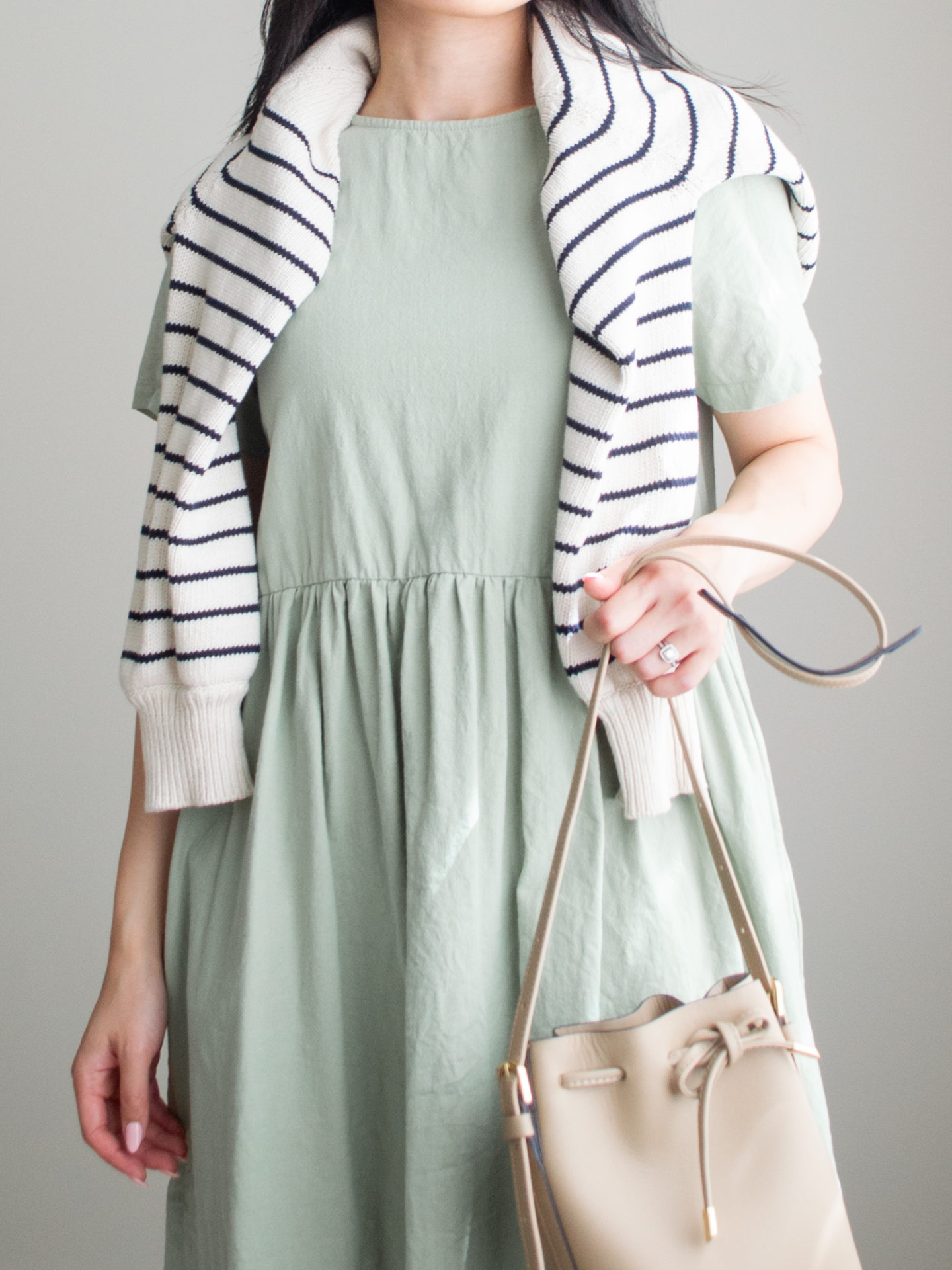 Close-up detailed outfit shot of Sharon of Her Simple Sole wearing a sage relaxed mini dress and a striped sweater over the shoulders, while holding a brown bucket bag | minimalist outfit ideas | baby doll dress outfit