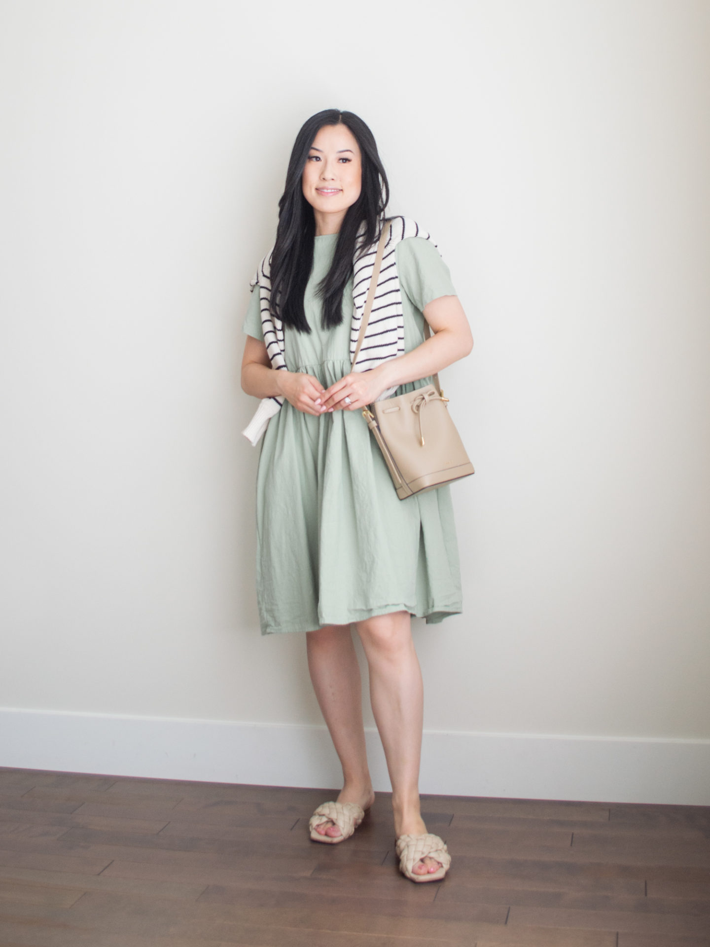 Sharon of Her Simple Sole is wearing items from her list of 10 summer wardrobe essentials, including a sage relaxed mini dress and a striped sweater over the shoulders, while holding a brown bucket bag | minimalist outfit ideas | baby doll dress outfit