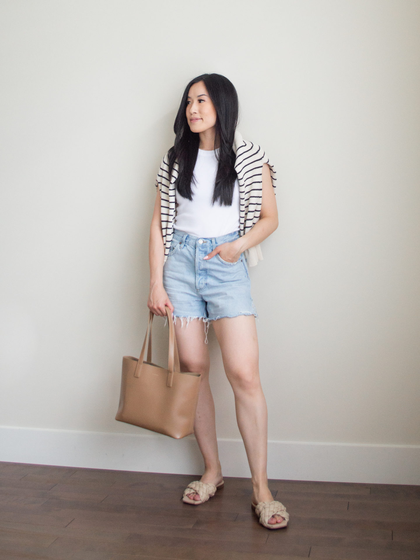 Sharon of Her Simple Sole is wearing items from her list of 10 summer wardrobe essentials, including a basic cotton tank top, a striped sweater over the shoulders, and relaxed denim shorts, while holding a brown tote bag | minimalist outfit ideas | tank top and denim shorts outfit