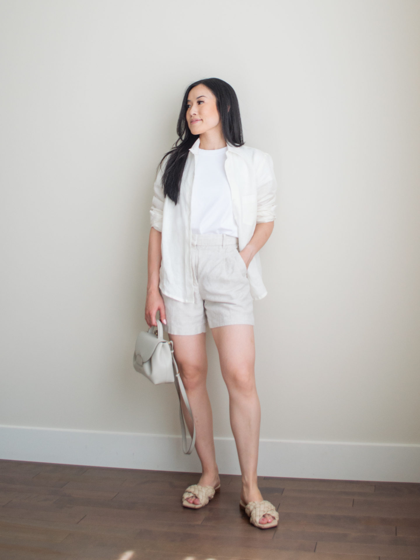 Sharon of Her Simple Sole is wearing a few items from her list of 10 summer wardrobe essentials, including a basic white t-shirt, a linen long-sleeve shirt, linen shorts, and criss-cross sandals, while holding a Polene bag | minimalist outfit ideas | light toned outfit