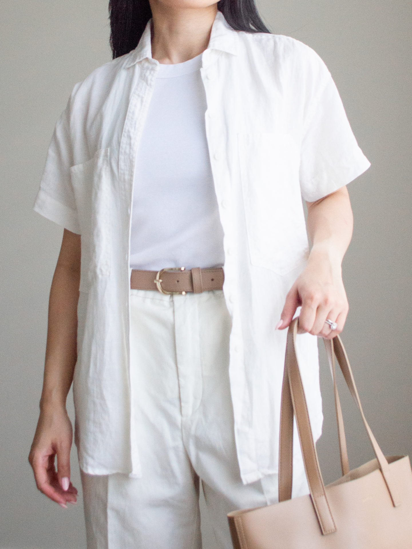 Close-up detailed outfit shot of Sharon of Her Simple Sole wearing a linen short sleeve button-up shirt, a basic white tank top underneath, linen pants, and a neutral belt, while holding a brown tote bag | minimalist outfit ideas | striped linen shirt and pants outfit