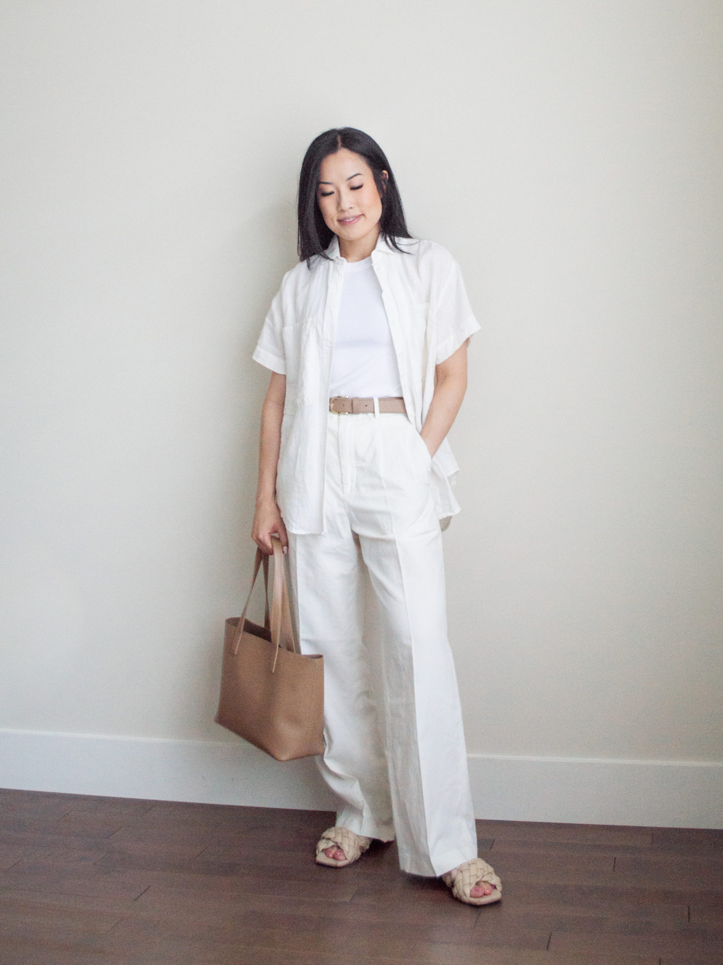 Sharon of Her Simple Sole is wearing items from her list of 10 summer wardrobe essentials, including a linen short sleeve button-up shirt, a basic white tank top underneath, linen pants, a neutral belt, and criss-cross sandals, while holding a brown tote bag | minimalist outfit ideas | striped linen shirt and pants outfit