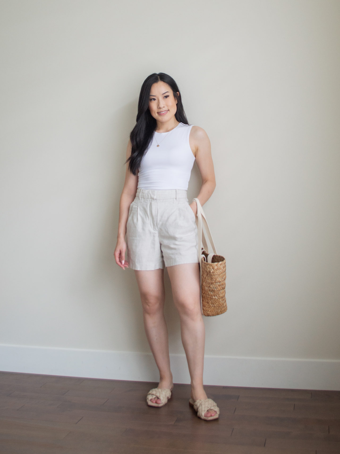 Sharon of Her Simple Sole is wearing an outfit using tips on how to elevate basic summer wardrobe essentials, including a seamless white bodysuit and linen blend shorts, while holding a brown basket bag and wearing criss cross braided sandals | basic summer wardrobe essentials