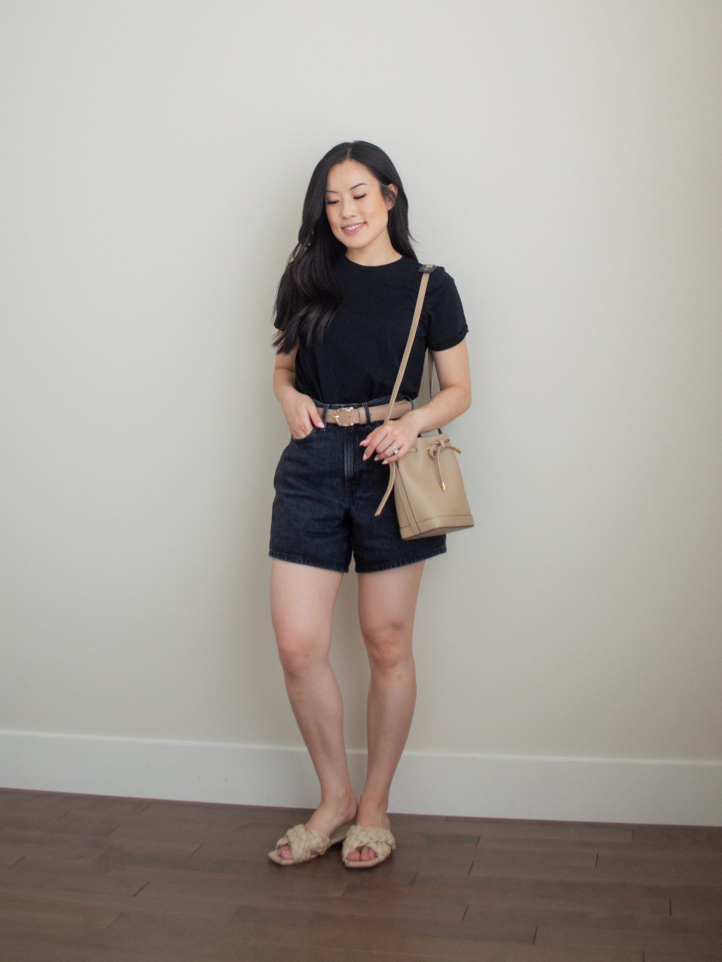 Sharon of Her Simple Sole is wearing an outfit using tips on how to elevate basic summer wardrobe essentials, featuring a basic black t-shirt, black jean shorts, and taupe belt, while holding a taupe bucket bag and wearing criss cross braided sandals | minimalist outfit ideas | basic summer wardrobe essentials