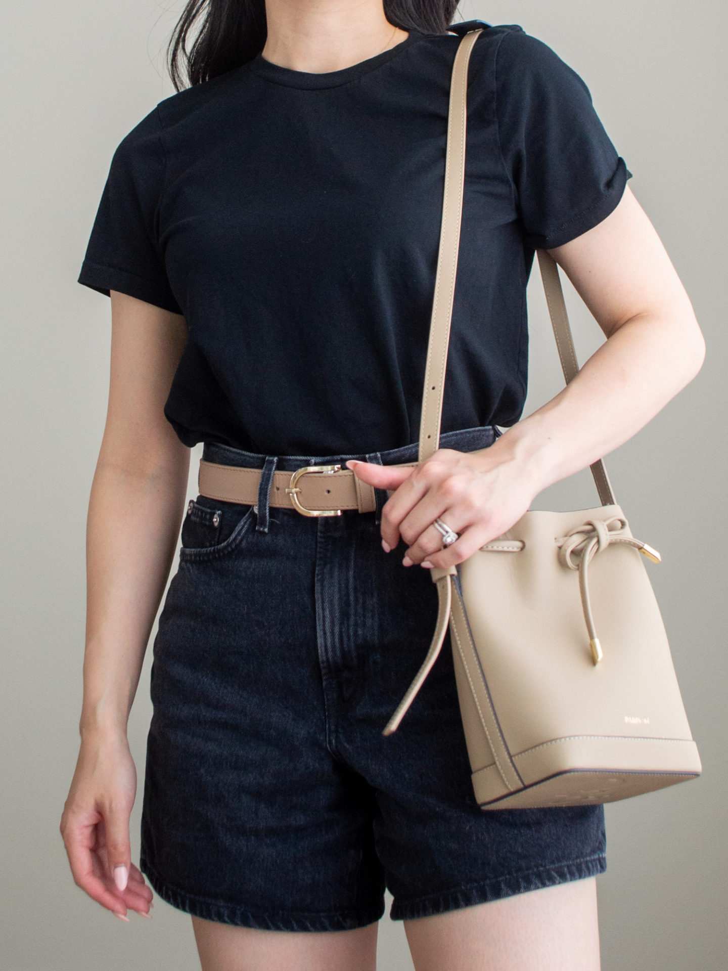 Close-up detailed outfit shot of Sharon of Her Simple Sole is wearing a basic black t-shirt, black jean shorts, and taupe belt, while holding a taupe bucket bag | minimalist outfit ideas | tshirt and denim shorts outfit