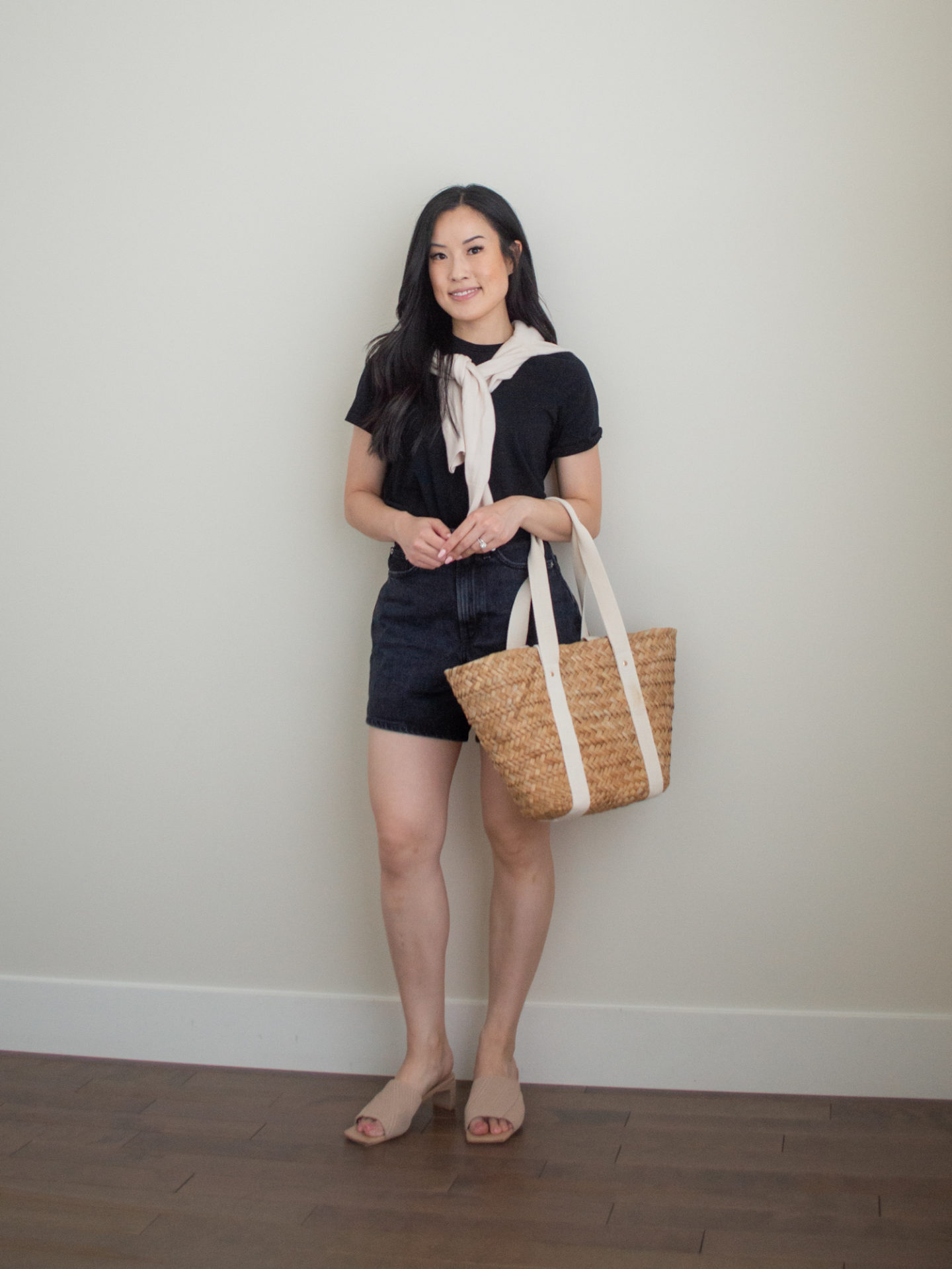 Sharon of Her Simple Sole is wearing an outfit using tips on how to elevate basic summer wardrobe essentials, featuring a knit sweater over the shoulders, a black t-shirt, and black jean shorts, while holding a brown straw bag and wearing neutral open toe mules | minimalist outfit ideas | basic summer wardrobe essentials