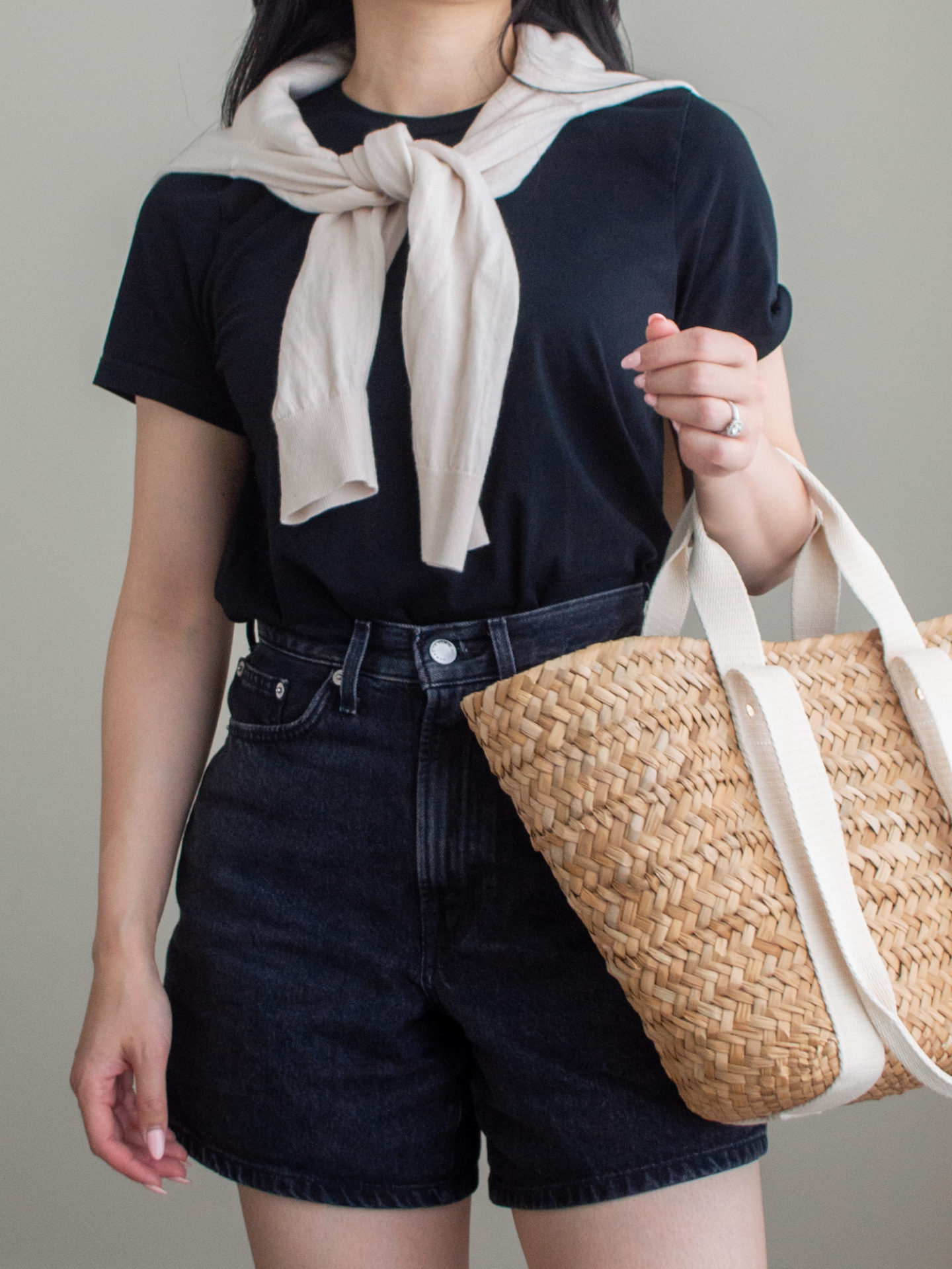 Close-up detailed outfit shot of Sharon of Her Simple Sole is wearing a knit sweater over the shoulders, a black t-shirt, and black jean shorts, while holding a brown straw bag | minimalist outfit ideas | sweater over shoulders outfit