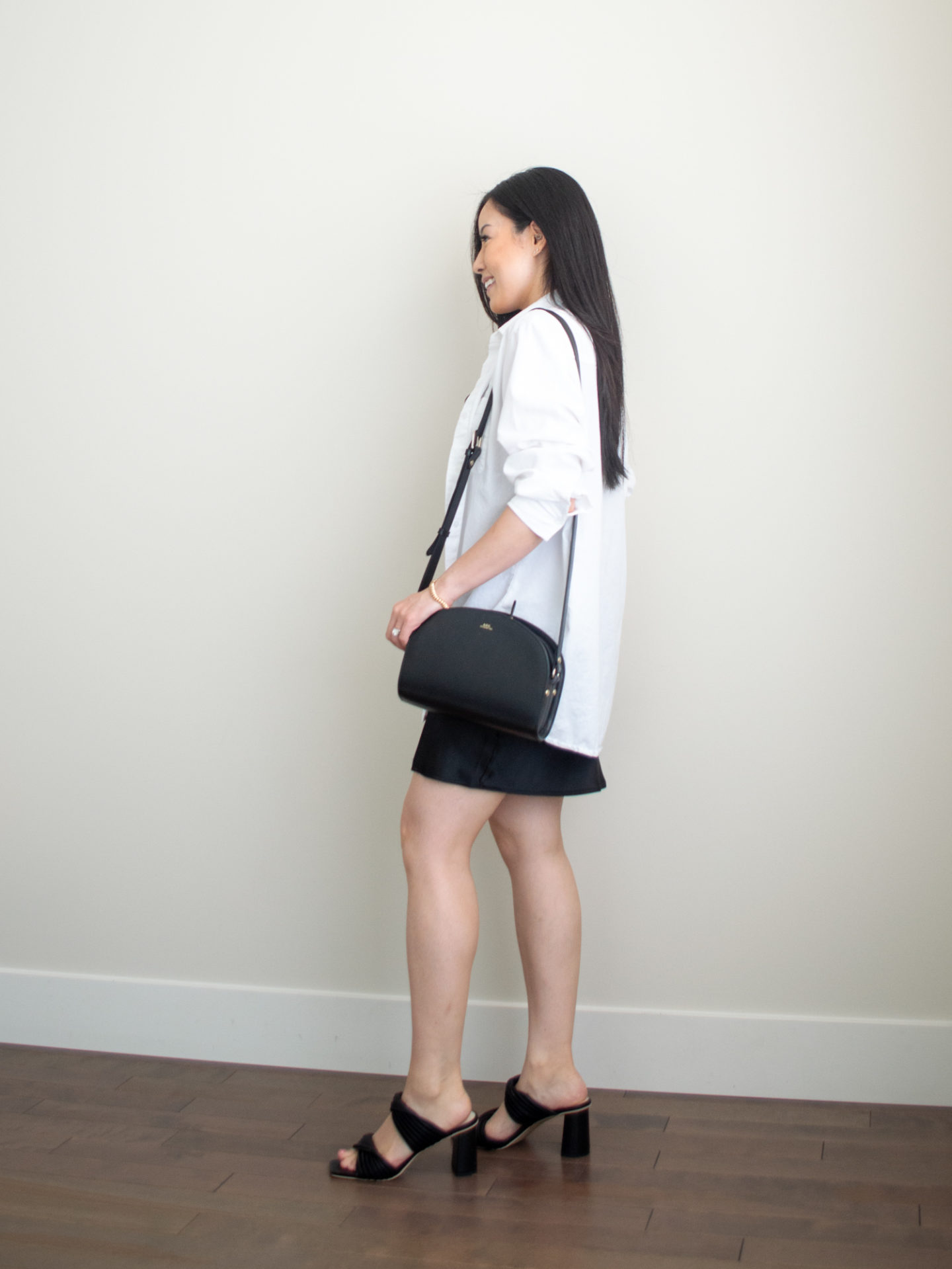 Sharon of Her Simple Sole is wearing an outfit featuring a black cowl neck mini slip dress, with an oversized white button-up shirt while holding a black crossbody bag and wearing block heeled sandals | minimalist outfit ideas | little black dress outfit