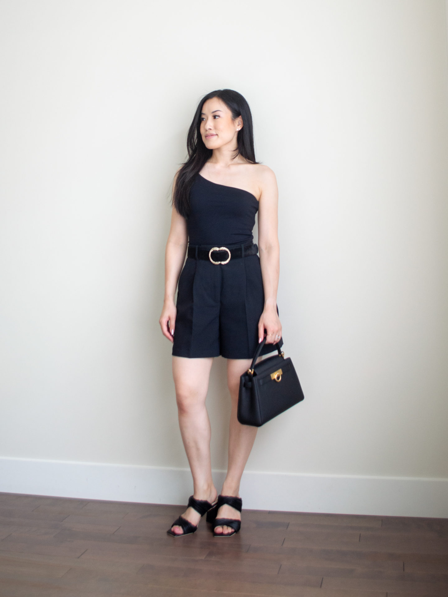 Sharon of Her Simple Sole is wearing an outfit from her 5 Simple Summer Date Night Outfits blog post featuring a black asymmetrical top, black tailored shorts, and a statement black belt, while holding a black top handle bag and wearing block heeled sandals | minimalist outfit ideas | summer date night outfits