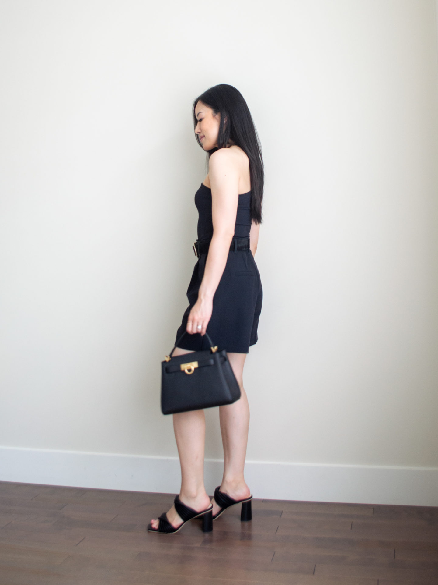 Sharon of Her Simple Sole is wearing a black asymmetrical top, black tailored shorts, and a statement black belt, while holding a black top handle bag | minimalist outfit ideas | black asymmetrical top |