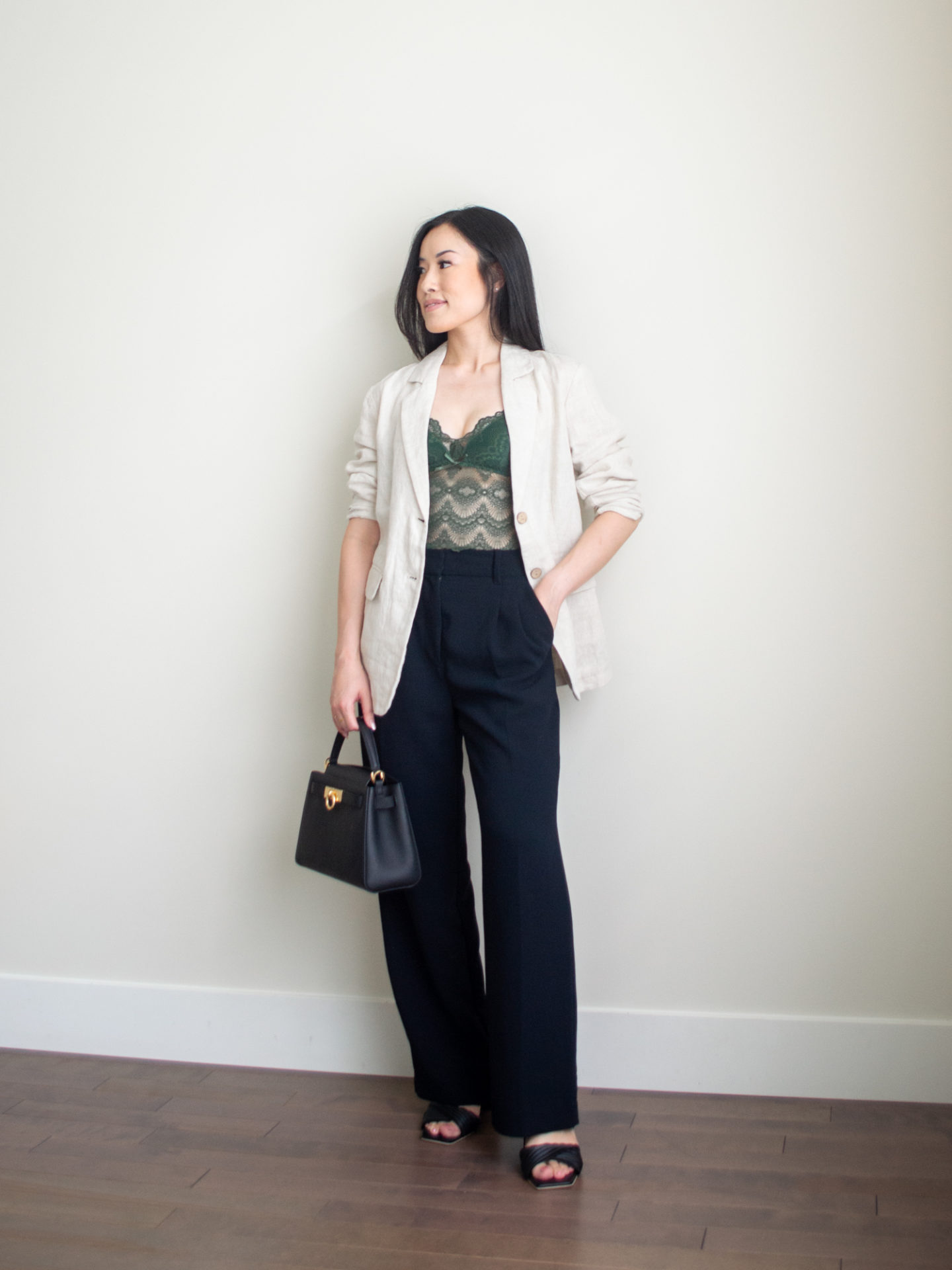 Sharon of Her Simple Sole is wearing an outfit from her 5 Simple Summer Date Night Outfits blog post featuring a green lace bustier, linen blazer, and black wide leg pants, while holding a black top handle bag and wearing block heeled sandals | minimalist outfit ideas