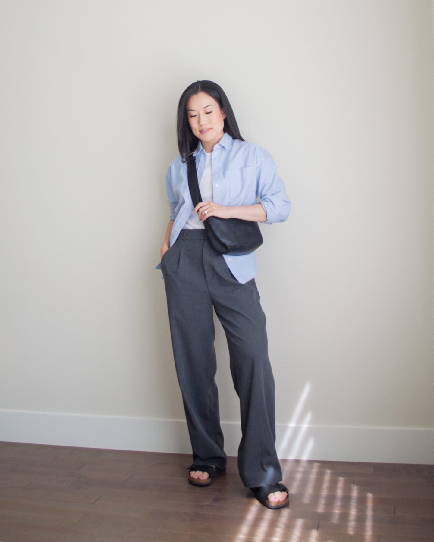 Sharon of Her Simple Sole is wearing an outfit from her June outfit roundup, including a white baby t-shirt, a blue relaxed oxford button up shirt, and grey wide leg pants, while holding a black slouchy bag and wearing birkenstock sandals | minimalist outfit ideas | smart casual outfit