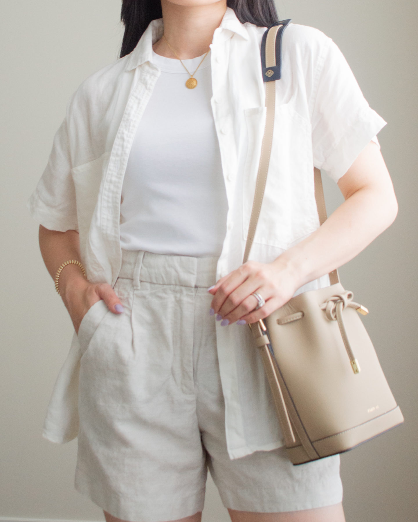 Close-up detailed outfit shot of Sharon of Her Simple Sole wearing a basic white tank top, a linen short sleeve button up shirt, and linen blend shorts, while holding a taupe bucket bag | minimalist outfit ideas | smart casual outfit