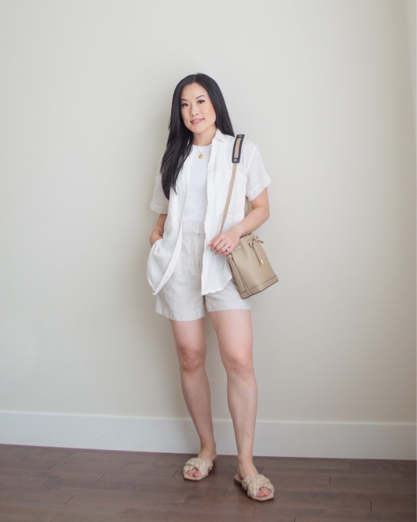 Sharon of Her Simple Sole is wearing a basic white tank top, a linen short sleeve button up shirt, and linen blend shorts, while holding a taupe bucket bag and wearing criss cross braided sandals | minimalist outfit ideas | summer outfit idea | linen and cotton outfit