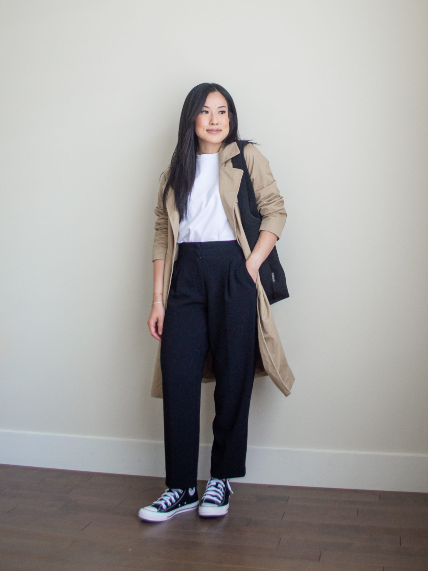 Sharon of Her Simple Sole is showcasing a t-shirt outfit for fall, featuring a simple white T-shirt, brown trench coat, and black cropped trousers, while holding a black tote bag and wearing a pair of black converse shoes | minimalist outfits | fall outfit ideas