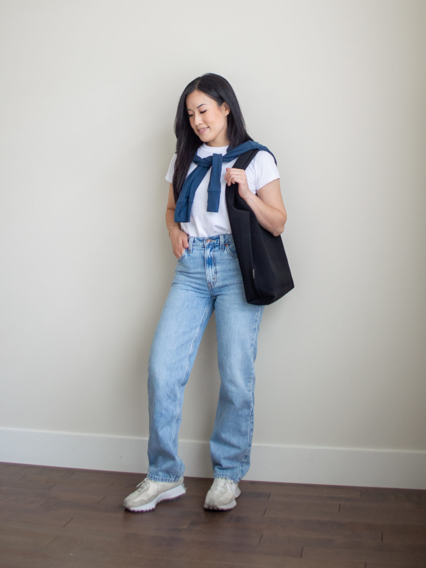 Sharon of Her Simple Sole is showcasing a t-shirt outfit for fall, featuring a simple white T-shirt, a blue sweatshirt tied over the shoulder, a pair of wide-leg jeans, a black tote bag and a pair of New Balance 327 sneakers | minimalist outfits | fall outfit ideas | minimalist