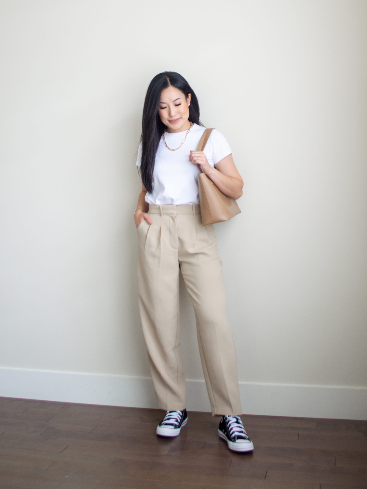 Sharon of Her Simple Sole is showcasing a t-shirt outfit for fall, featuring a simple white T-shirt, a chunky gold chain necklace, camel coloured tailored trousers, a caramel mini tote bag and a pair of black converse sneakers | minimalist outfits | fall outfit ideas