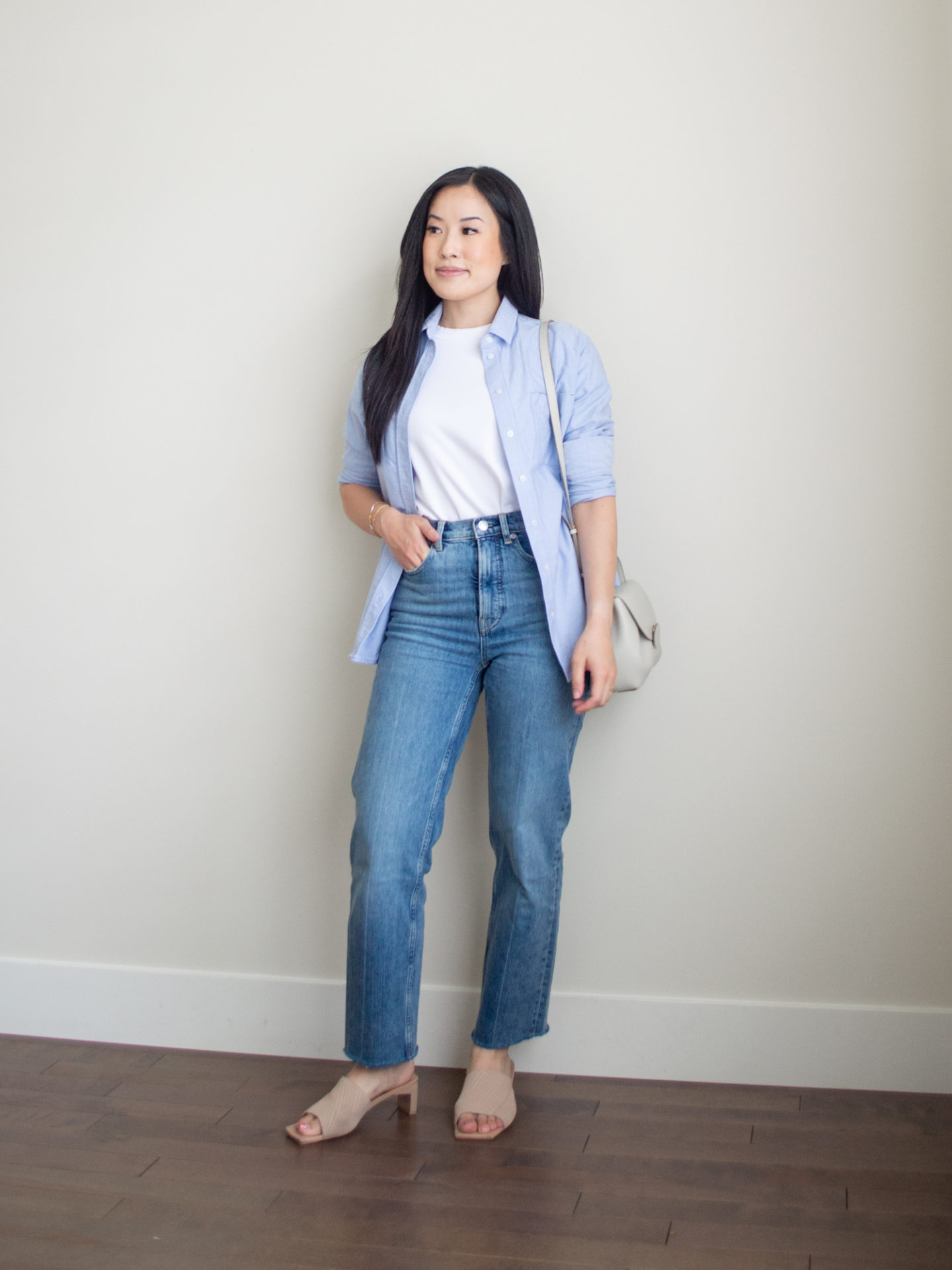 Sharon of Her Simple Sole is showcasing a t-shirt outfit for fall, featuring a simple white T-shirt, a blue relaxed button up shirt, a pair of blue straight leg jeans, an off-white crossbody bag and a pair of neutral open toe mules | minimalist outfits | fall outfit ideas