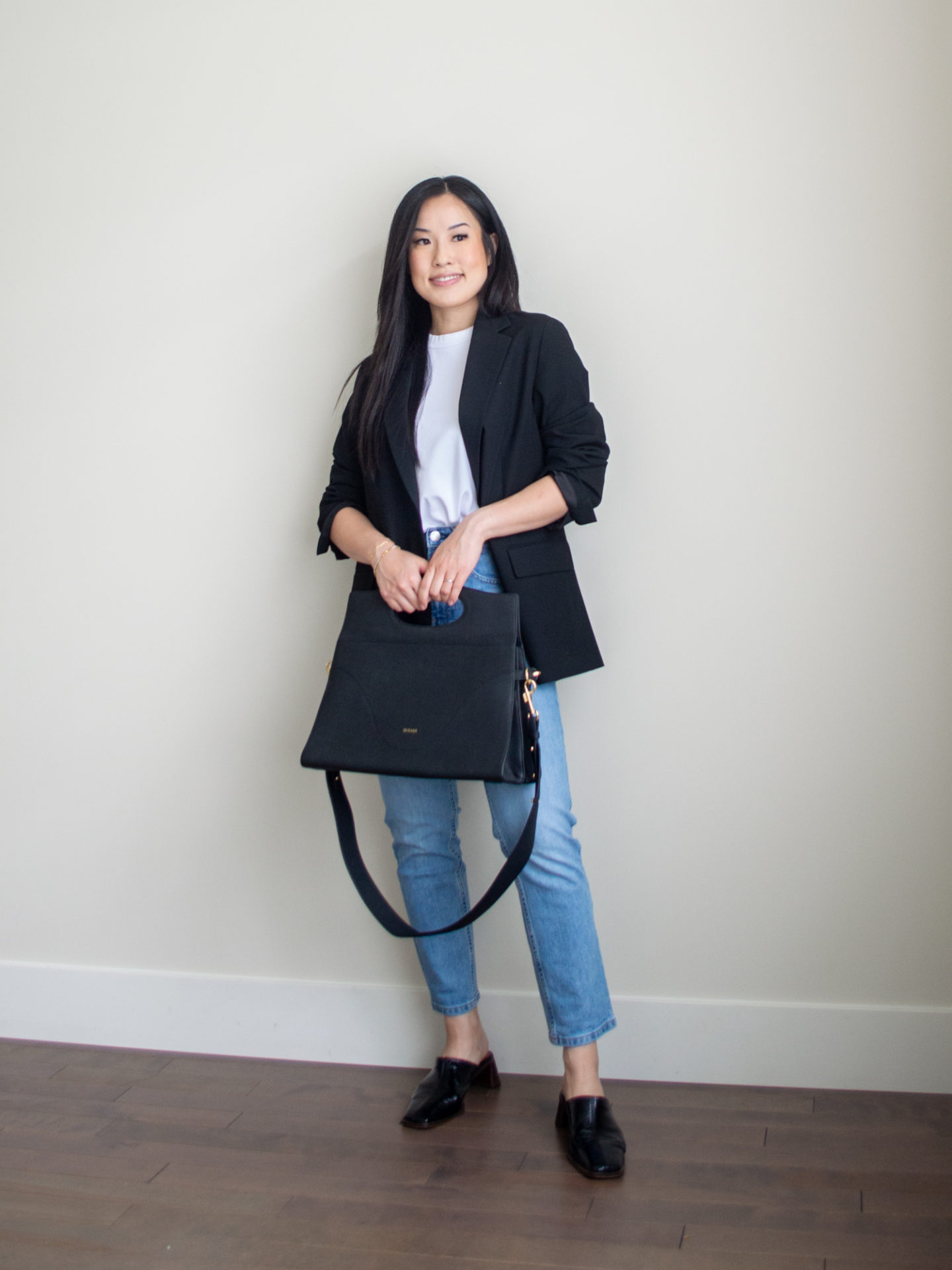 Sharon of Her Simple Sole is showcasing a t-shirt outfit for fall, featuring a simple white T-shirt, a black blazer, blue straight fit jeans, a black briefcase-style bag and black heeled mules | minimalist outfits | fall outfit ideas