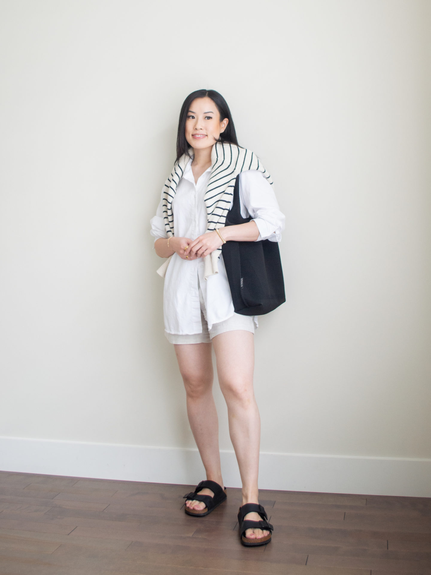 Sharon of Her Simple Sole is showcasing an outfit from her July outfit roundup, featuring an an oversized white button-up shirt, linen shorts, and a striped sweater over the shoulders while wearing Birkenstock sandals and black tote bag | minimalist outfits | summer outfit ideas | oversized button up shirt outfit ideas