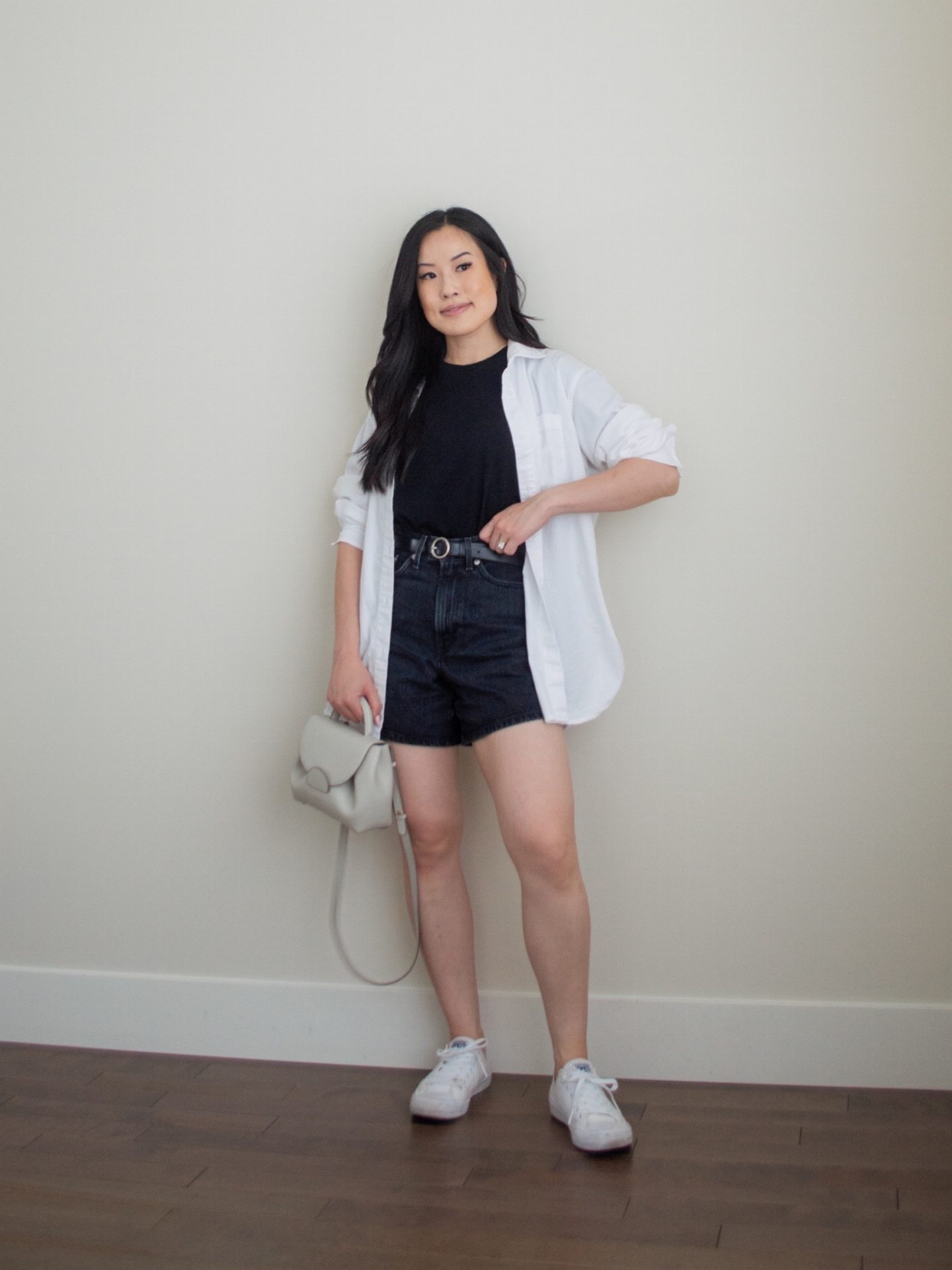 Sharon of Her Simple Sole is showcasing an outfit from her July outfit roundup, featuring a basic black t-shirt, black jean shorts, an oversized white button-up shirt, white sneakers, and a black belt while holding an off-white crossbody bag | minimalist outfits | summer outfit ideas | button up shirt outfit ideas