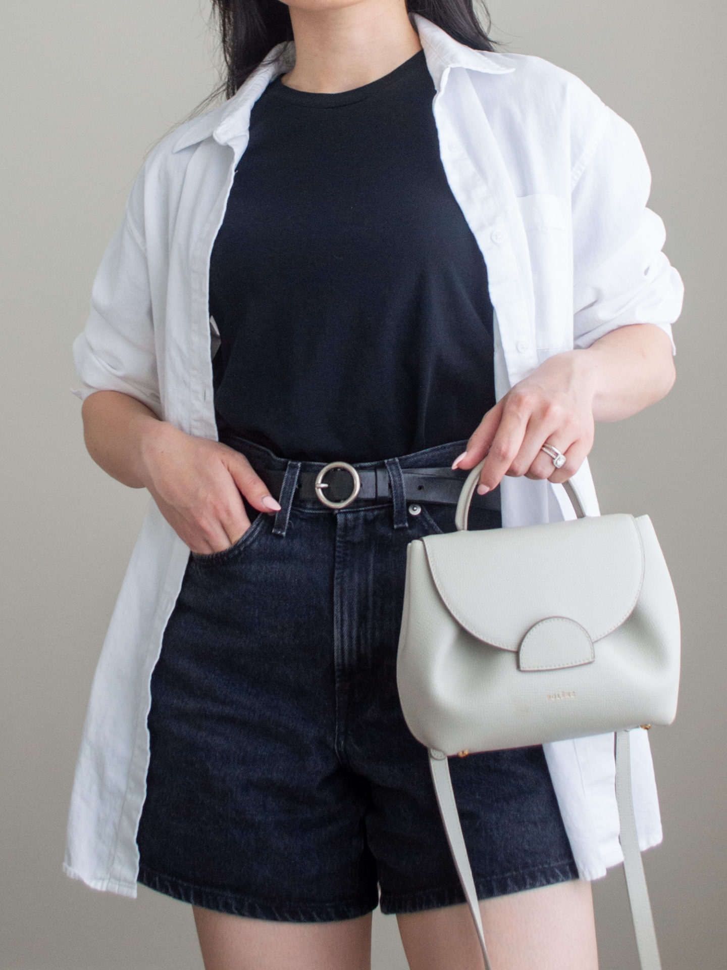 Close-up detailed outfit shot of Sharon of Her Simple Sole wearing a basic black t-shirt, black jean shorts, an oversized white button-up shirt, and a black belt while holding an off-white crossbody bag | minimalist outfits | summer outfit ideas | button up shirt outfit ideas