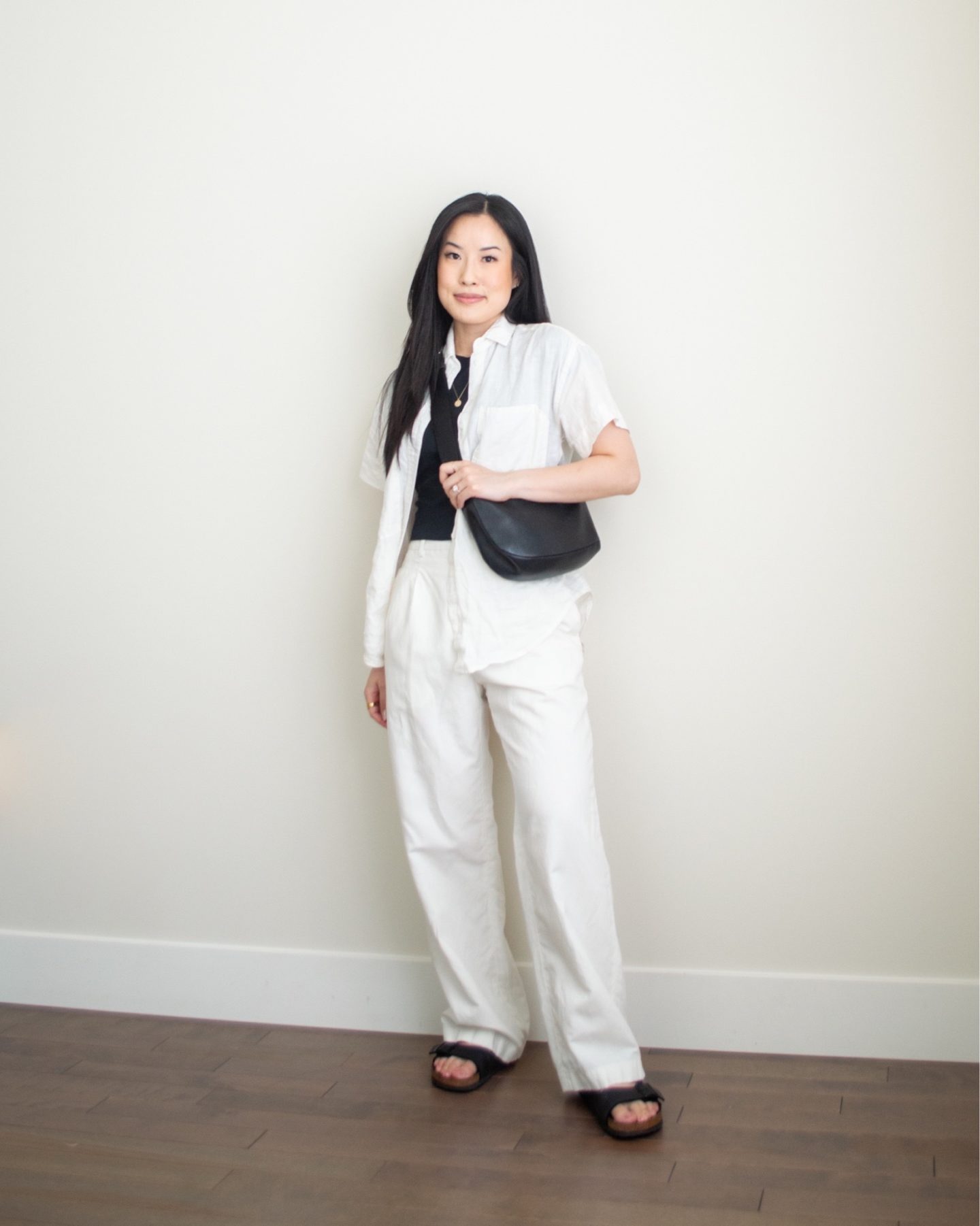 Sharon of Her Simple Sole is showcasing an outfit from her July outfit roundup, featuring a basic black T-shirt, a linen short sleeve button up shirt, cream wide leg linen pants, along with a black slouchy bag while wearing birkenstock sandals | minimalist outfits ideas | summer outfit ideas | linen outfit