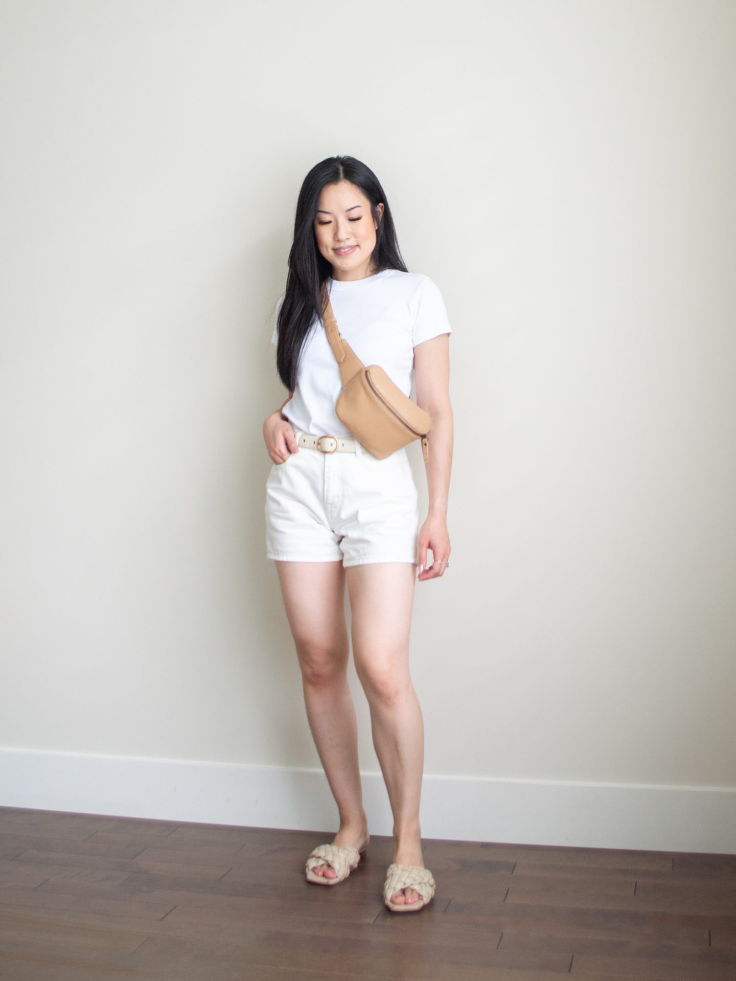 Sharon of Her Simple Sole is showcasing an outfit from her July outfit roundup, featuring a basic white T-shirt, white denim shorts, a cream belt, braided sandals and a tan belt bag | minimalist outfits ideas | summer outfit ideas | simple shirt