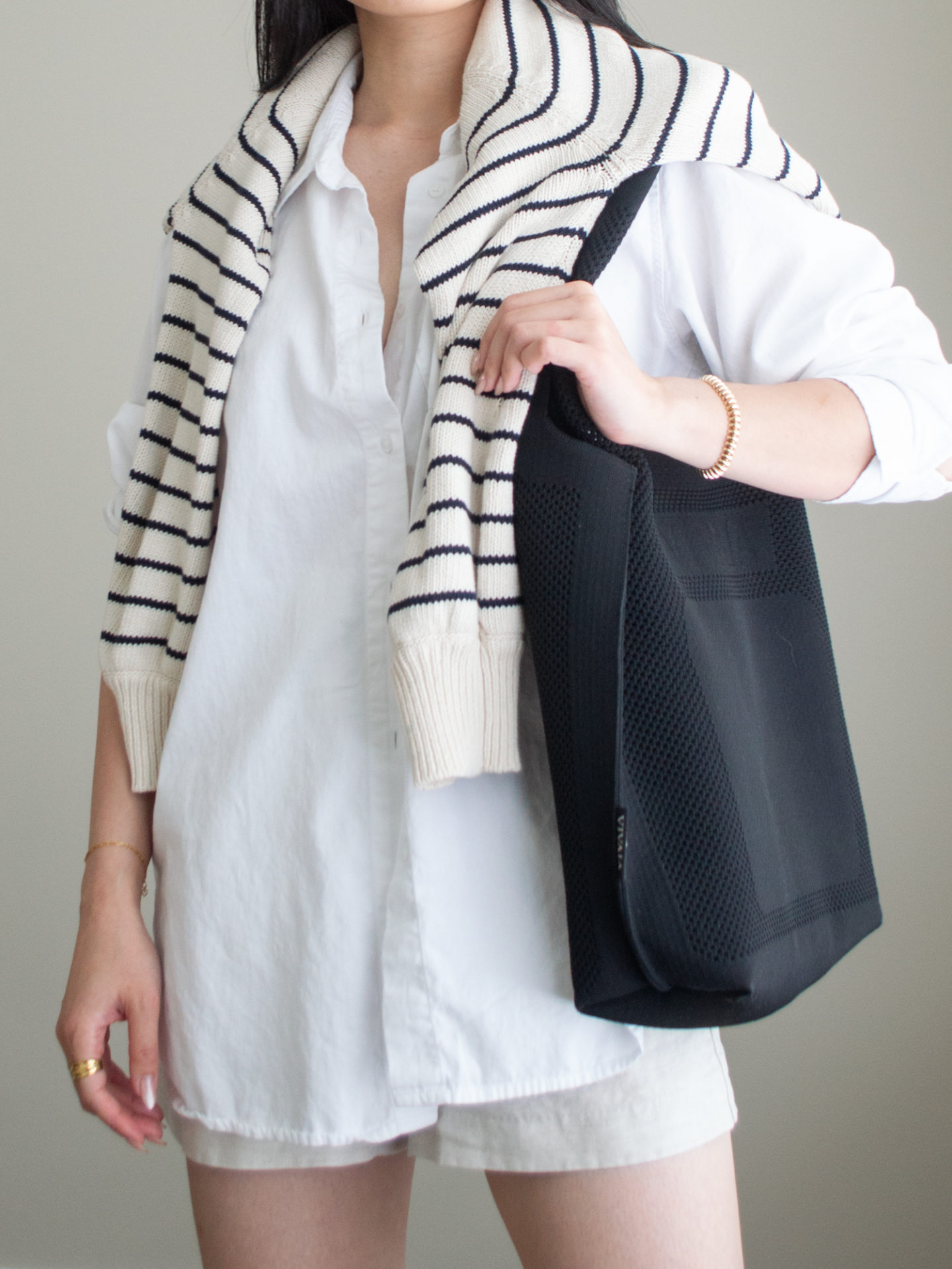 Close-up detailed outfit shot of Sharon of Her Simple Sole wearing an oversized white button-up shirt, linen shorts, and a striped sweater over the shoulders while holding a black tote bag | minimalist outfits | summer outfit ideas | oversized button up shirt outfit ideas