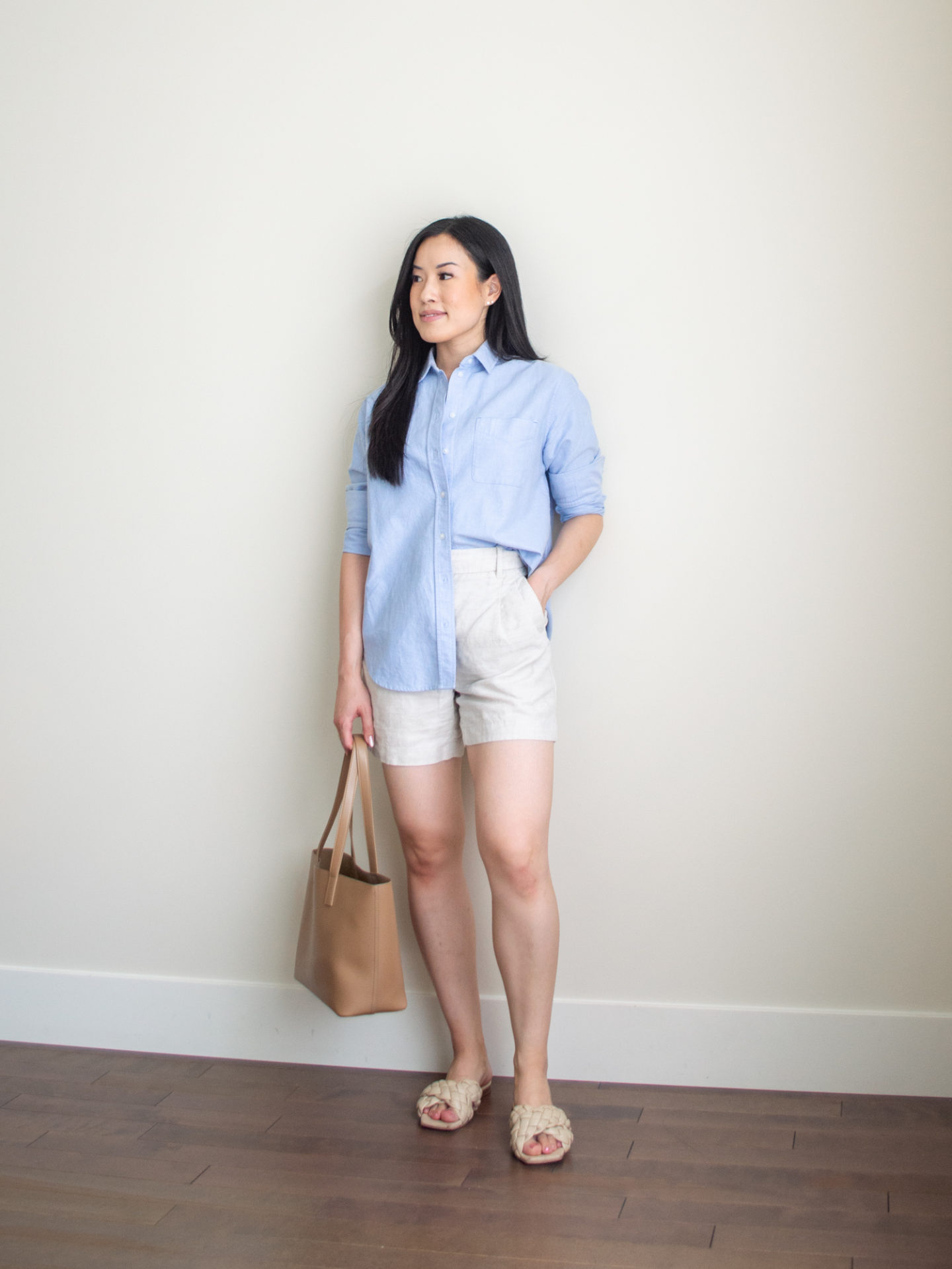 Sharon of Her Simple Sole is showcasing an outfit from her July outfit roundup, featuring a blue relaxed oxford button up shirt, linen shorts while wearing crisscross braided sandals and holding a brown tote bag | minimalist outfits | summer outfit ideas | blue button up shirt outfit