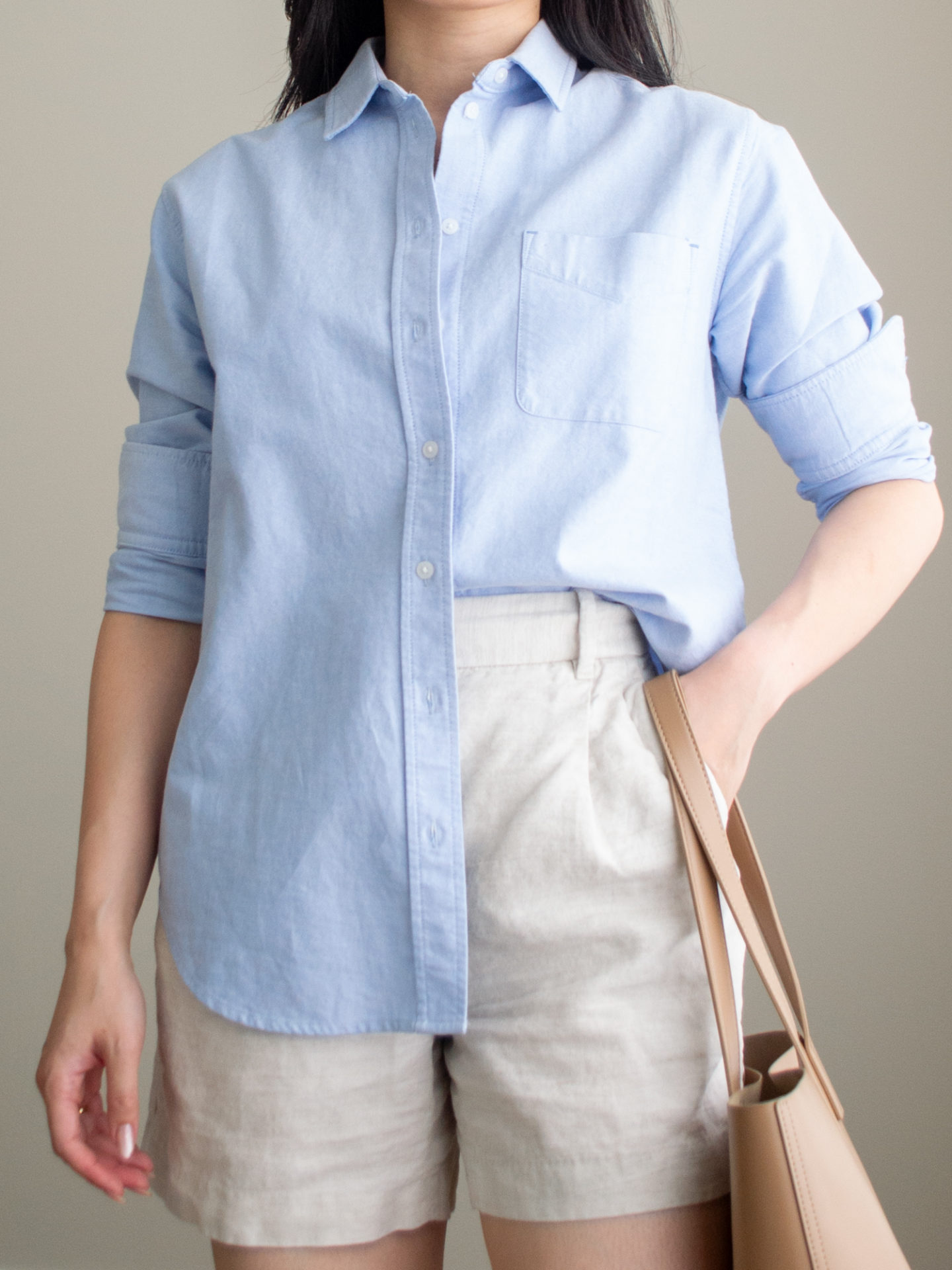 Close-up detailed outfit shot of Sharon of Her Simple Sole wearing a blue relaxed oxford button up shirt and linen shorts while holding a brown tote bag | minimalist outfits | summer outfit ideas | blue button up shirt outfit