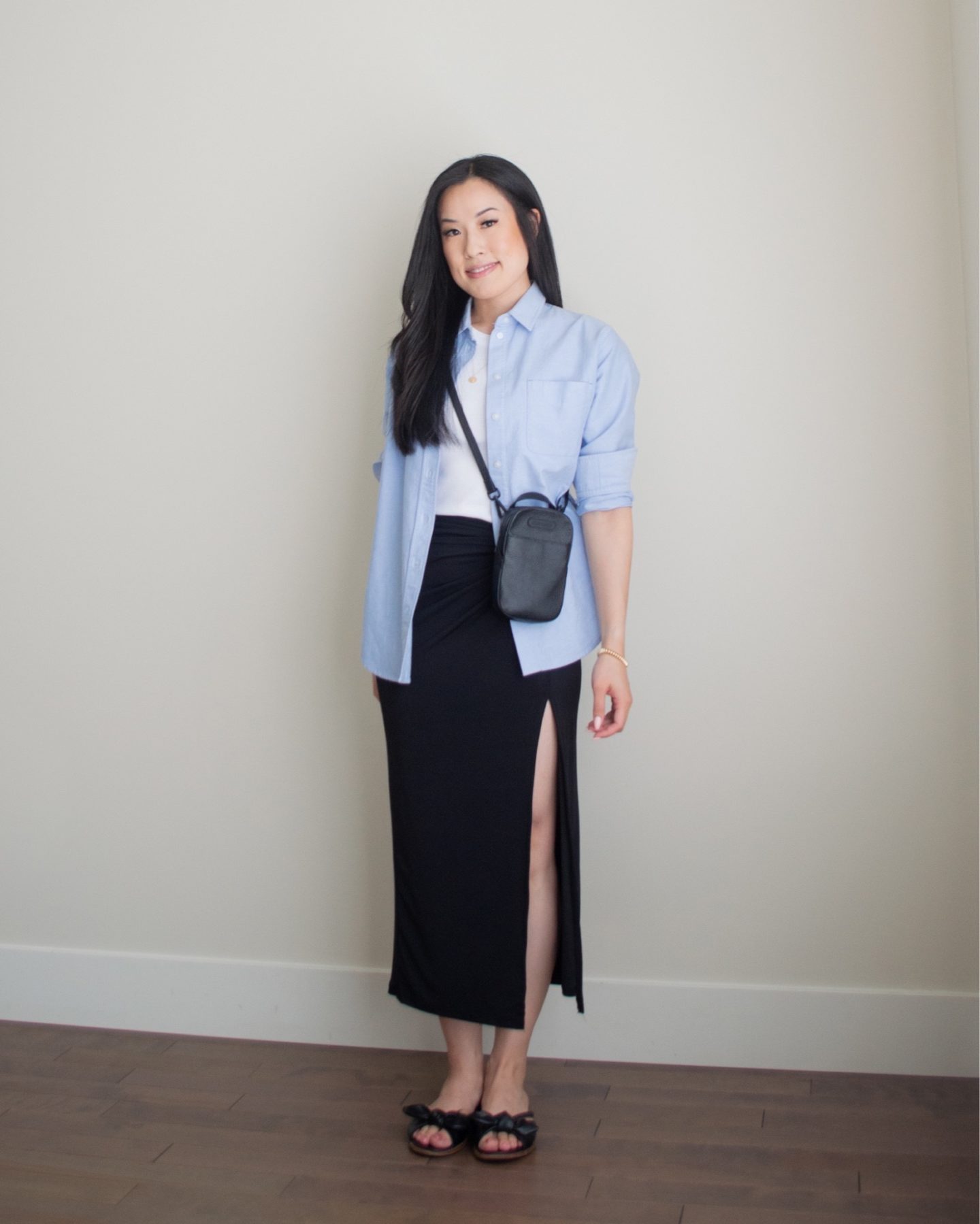 Sharon of Her Simple Sole is showcasing an outfit from her July outfit roundup, featuring a basic cotton tank top, a blue relaxed oxford button up shirt, a black midi skirt, a black crossbody phone bag, and a pair of black bow sandals | minimalist outfits | summer outfit ideas | button up shirt outfit