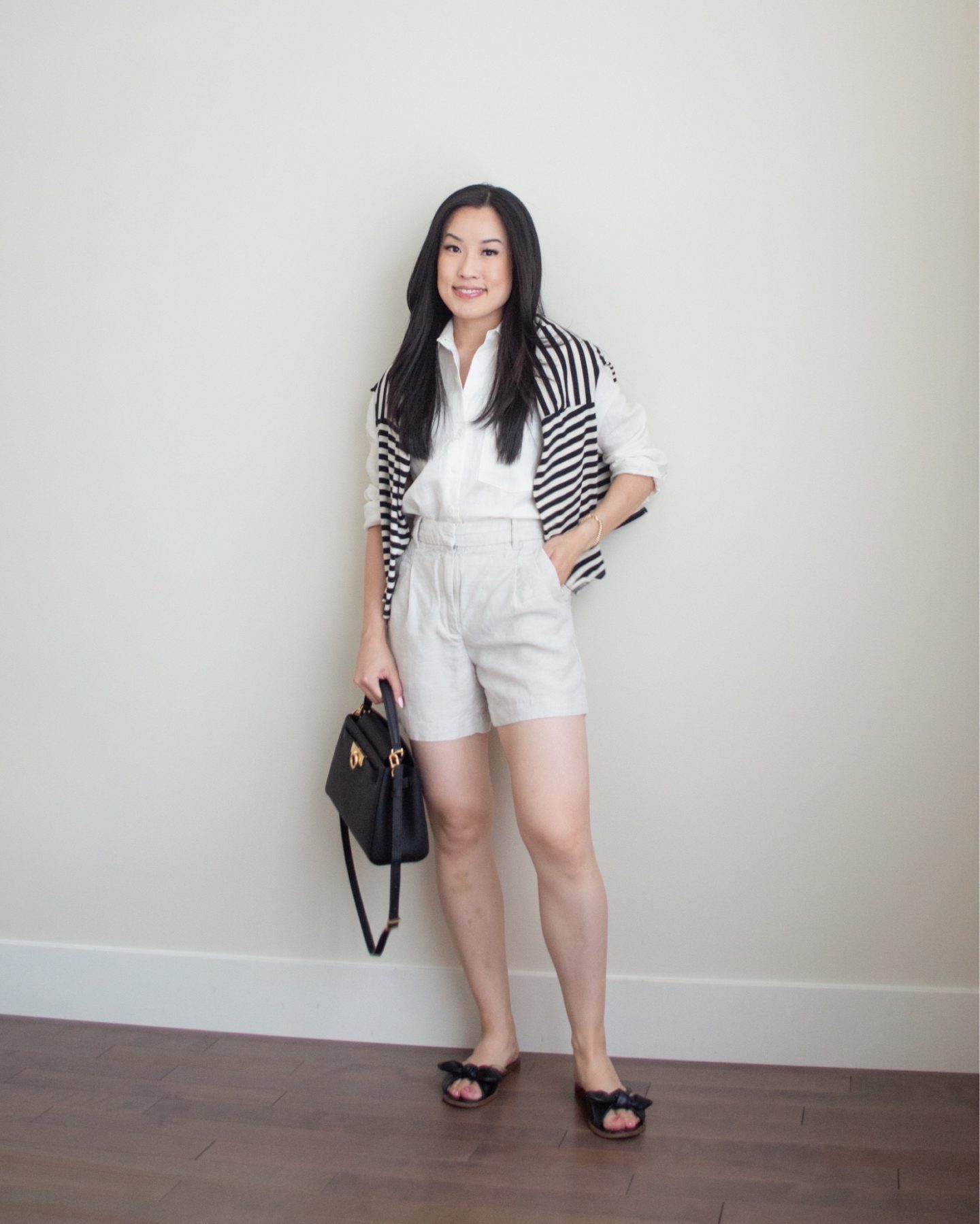 Sharon of Her Simple Sole is showcasing an outfit from her July outfit roundup, featuring a white linen button up shirt, a striped sweater over the shoulders along with linen shorts while holding a black top handle bag | minimalist outfits | summer outfit ideas | linen shirt outfit ideas