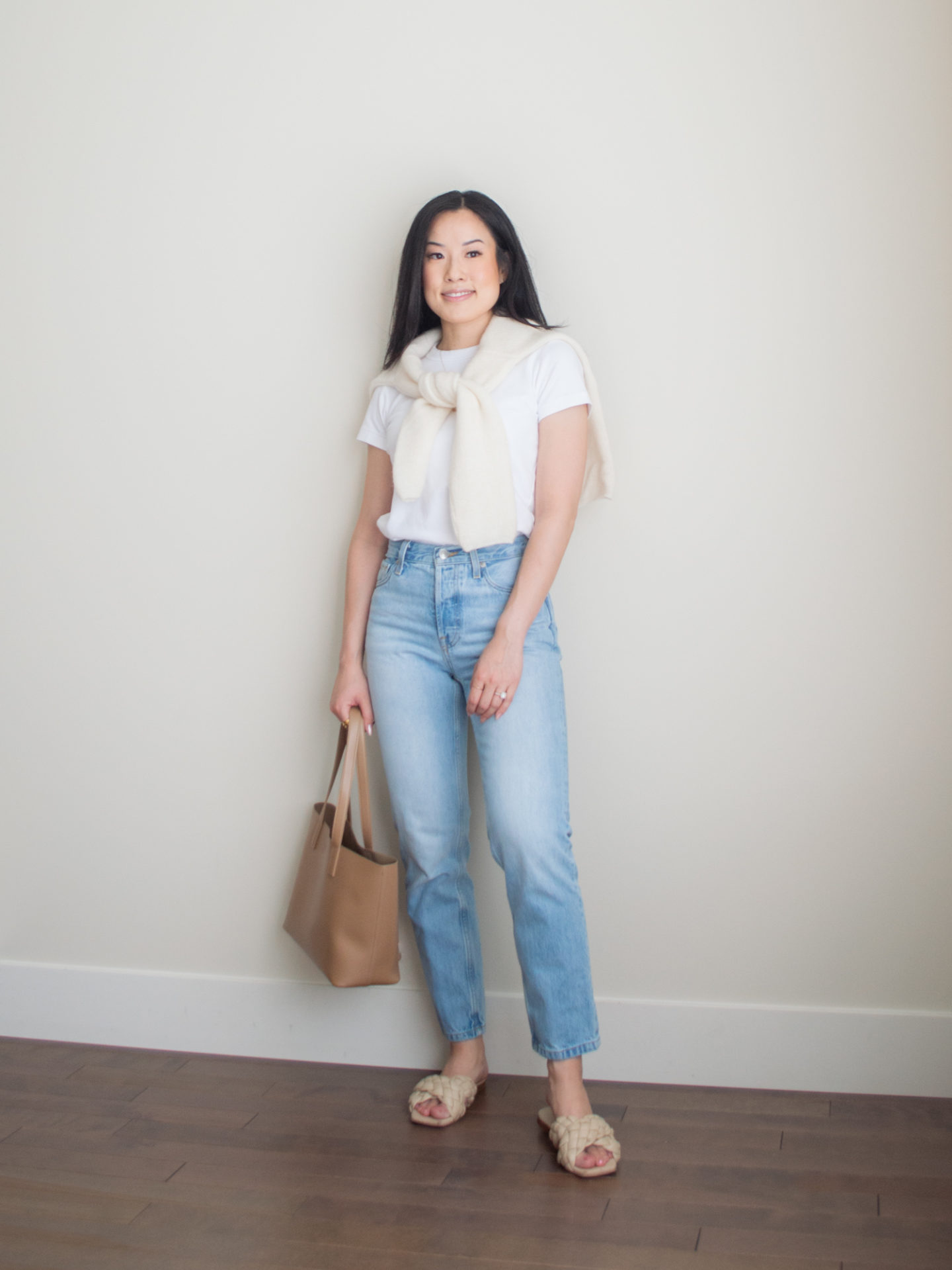 Sharon of Her Simple Sole is showcasing a outfit from her August outfit roundup, featuring a basic white T-Shirt, blue straight leg jeans, a wool cardigan over the shoulders, a caramel mini tote bag, and criss-cross cream sandals | minimalist outfits | simple outfit ideas