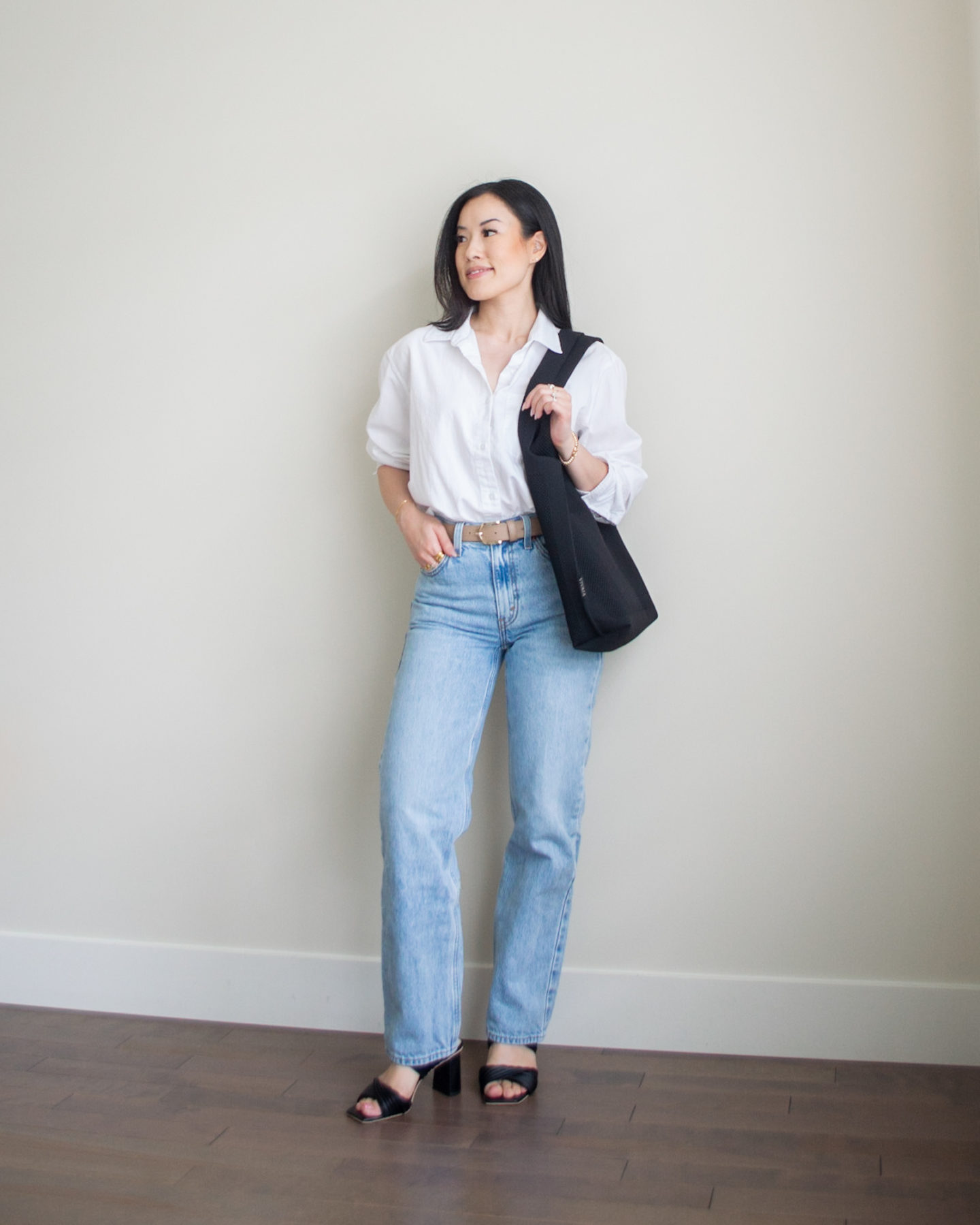 Sharon of Her Simple Sole is showcasing a outfit from her August outfit roundup, featuring a white oversized button up shirt, a pair of blue relaxed jeans, a black tote bag, a neutral toned belt, and a pair of black heeled sandals | minimalist outfits | simple outfit ideas