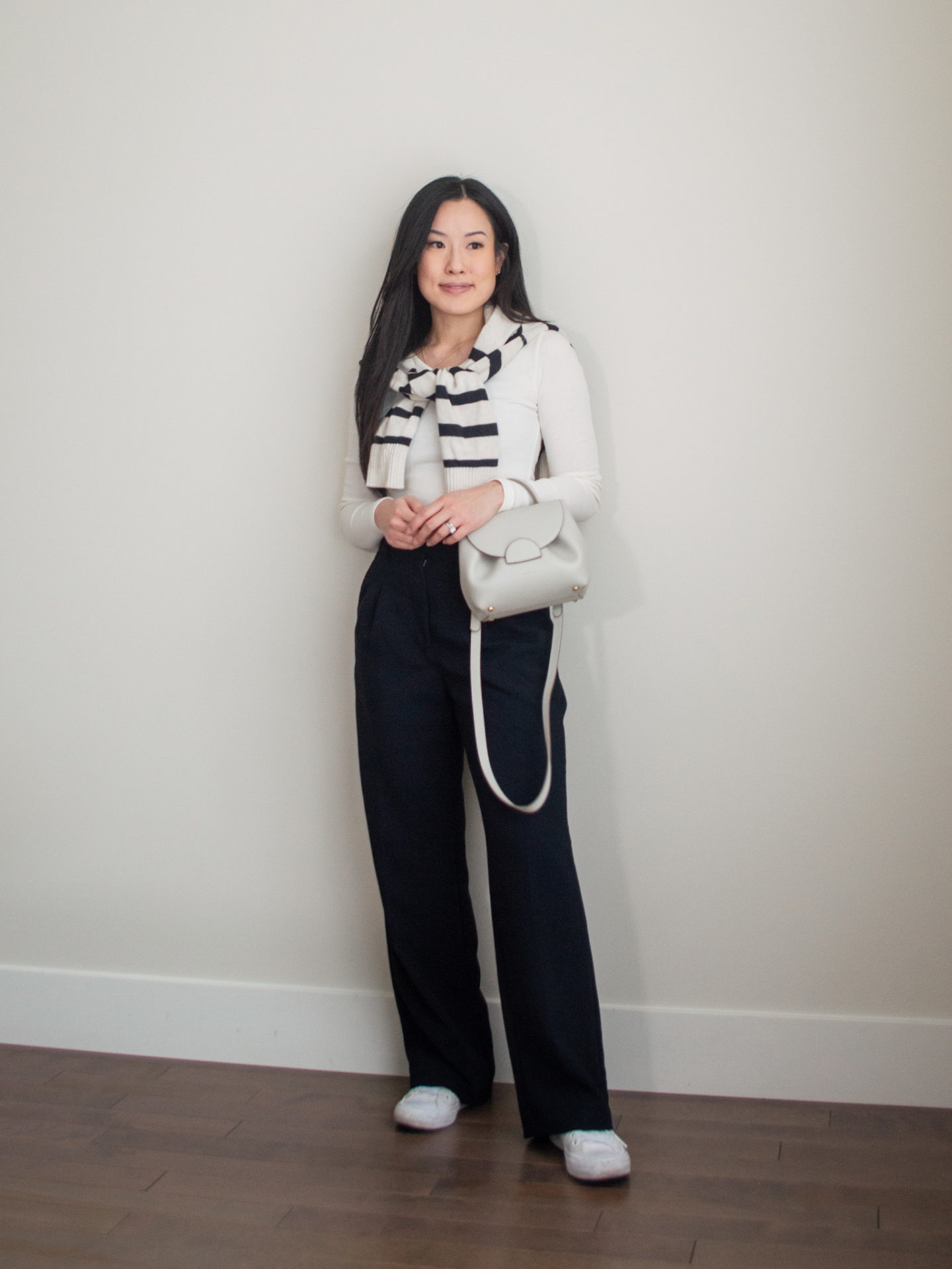 Sharon of Her Simple Sole is showcasing an outfit inspired by Parisian style, featuring a basic off-white long sleeve top, striped wool cardigan over the shoulder, black wide-leg pants, an off-white crossbody bag and white sneakers | minimalist outfits | fall outfit ideas | simple outfit ideas